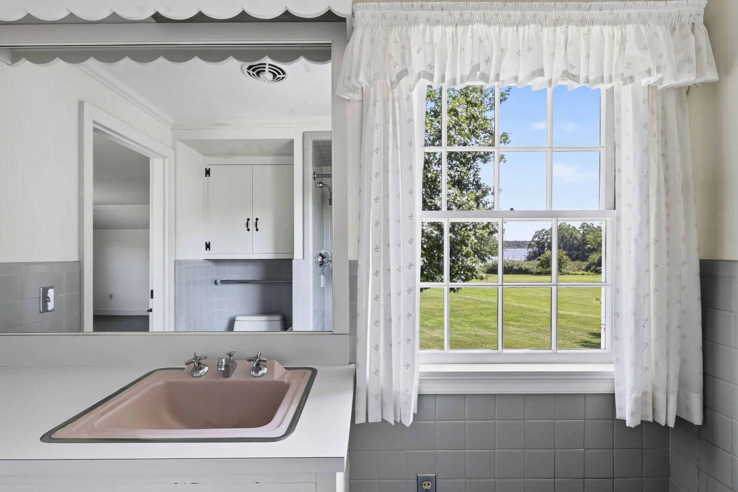 A bathroom with a view of a green landscape and water outside a large window, featuring a pink bathtub, white curtains, and a sink with faucets.