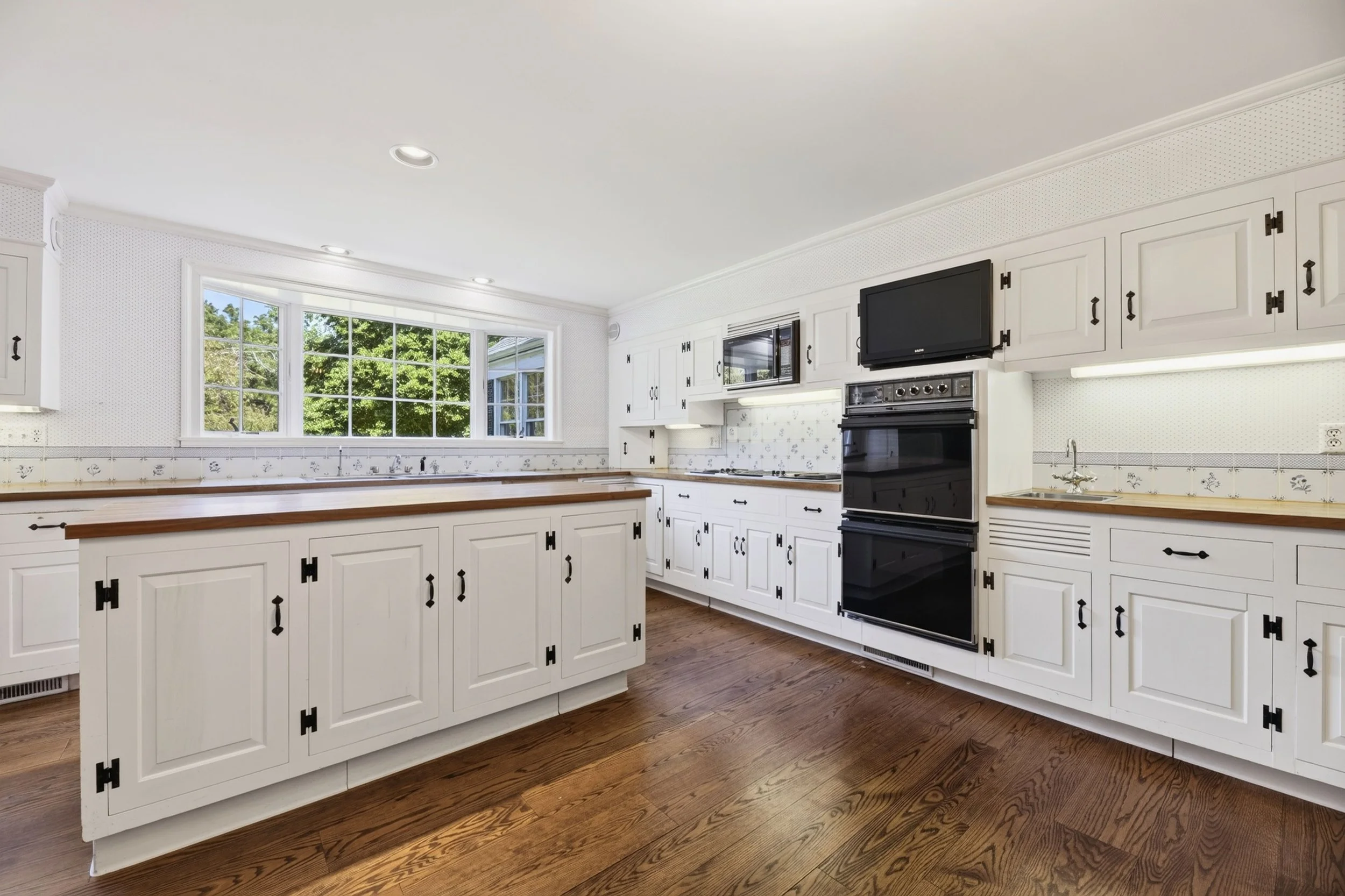 A spacious kitchen with white cabinets, wooden countertops, a large window, and a black oven and microwave. Hardwood floor and recessed ceiling lights.