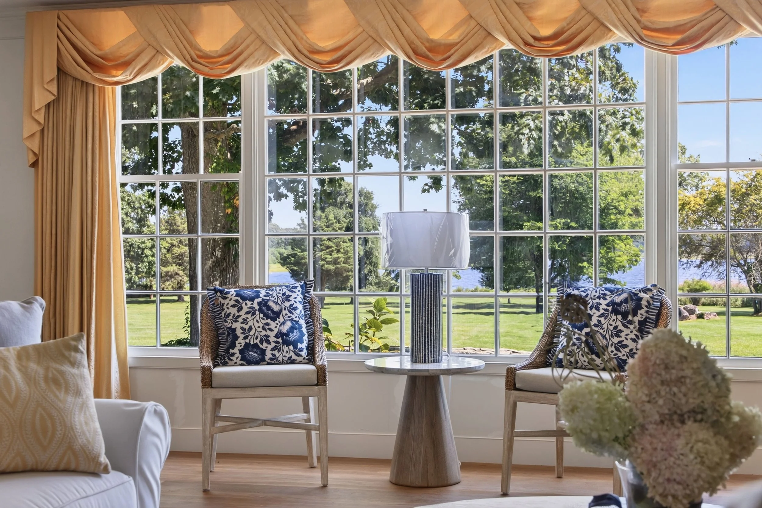 Living room interior with large window, orange draped curtains, wicker chairs with blue floral cushions, white sofa with yellow cushion, white round table with a striped lamp, and flowers in the foreground, overlooking a grassy yard and trees outside
