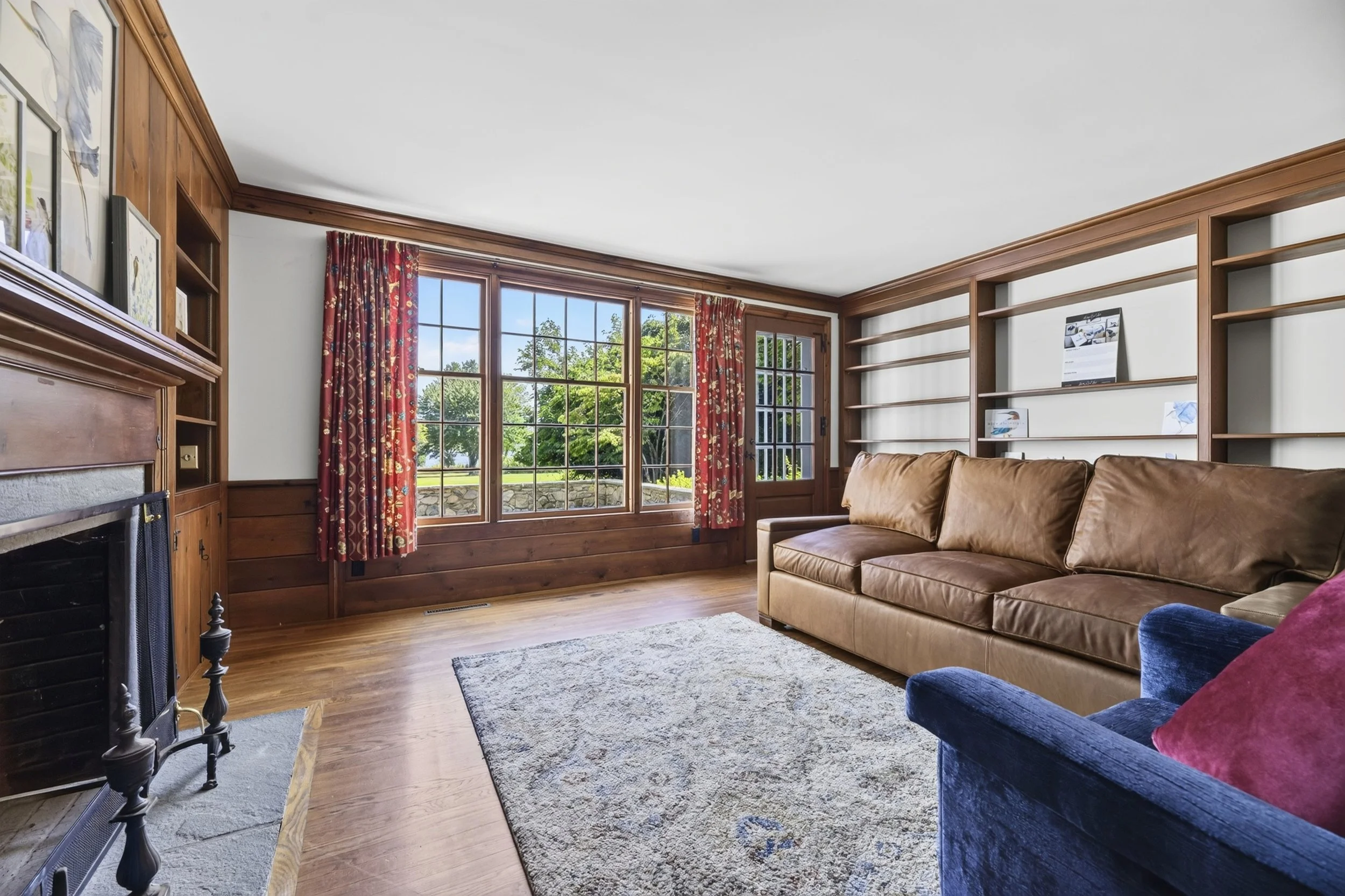 Living room with large window, red floral curtains, wooden window frame, brown leather sofa, blue velvet armchair with a red pillow, built-in wooden shelves, fireplace, and a white rug.