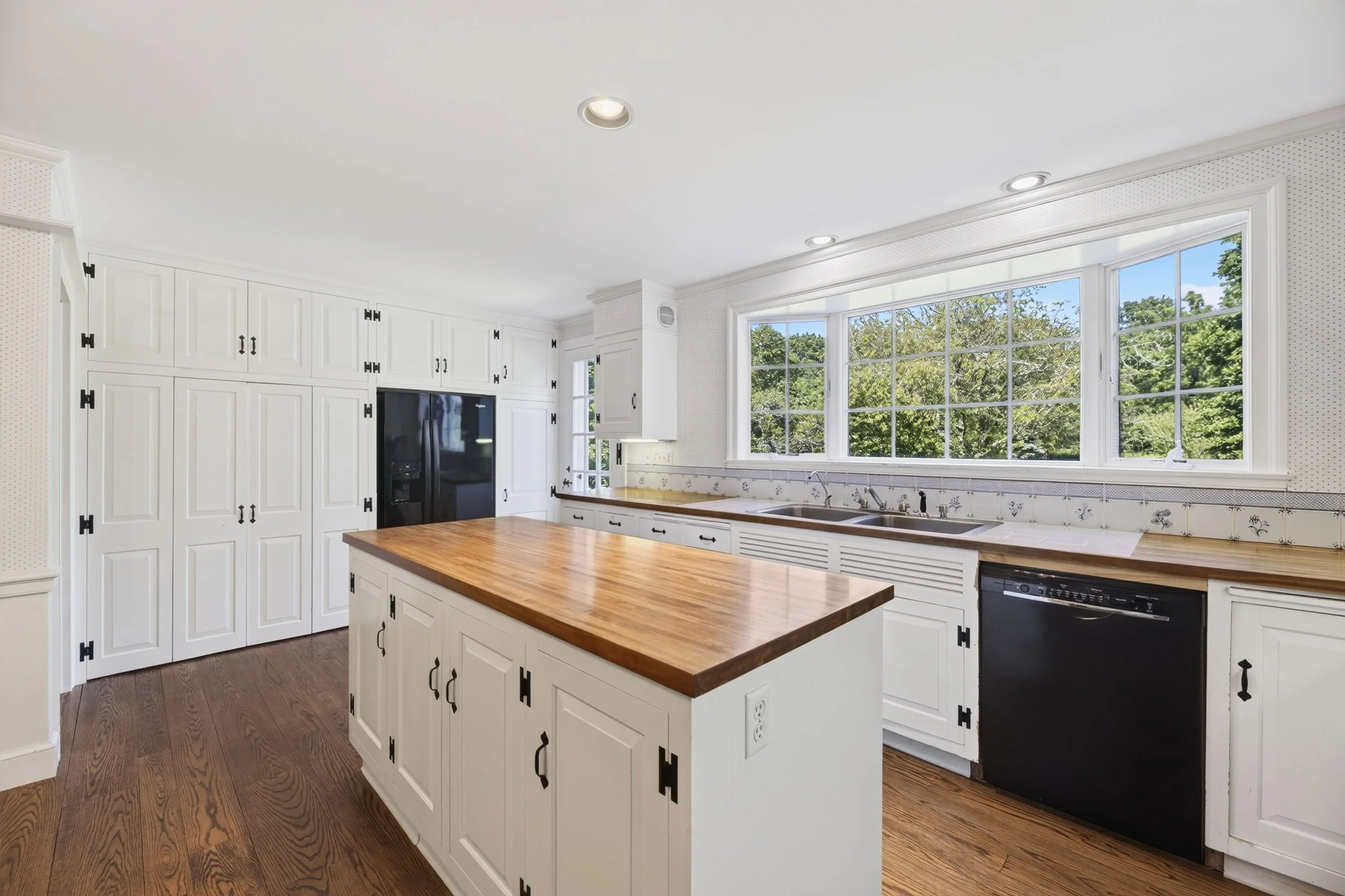Bright kitchen with white cabinets, wooden countertops, large window with green trees outside, and hardwood floors.