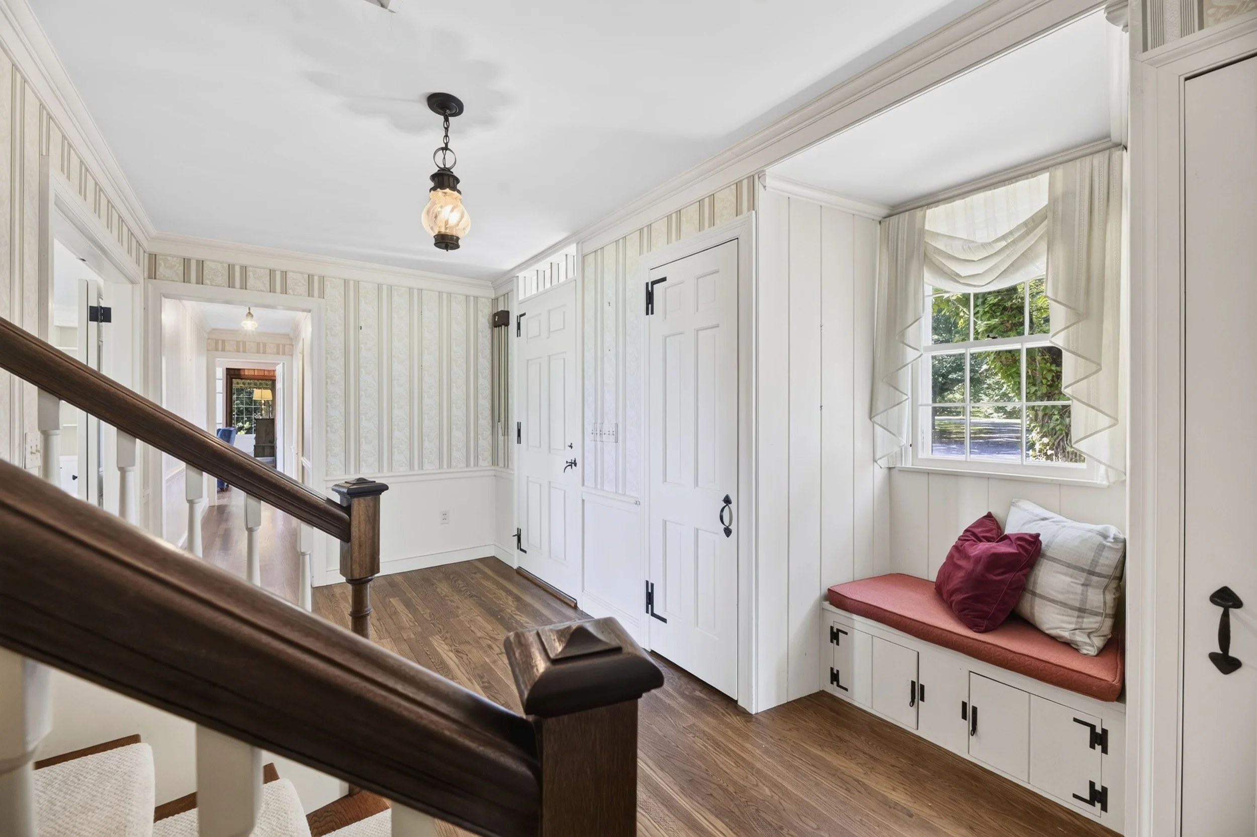 Interior of a house with a stairway, striped wallpaper, a window with curtains, and a built-in window seat with pillows.