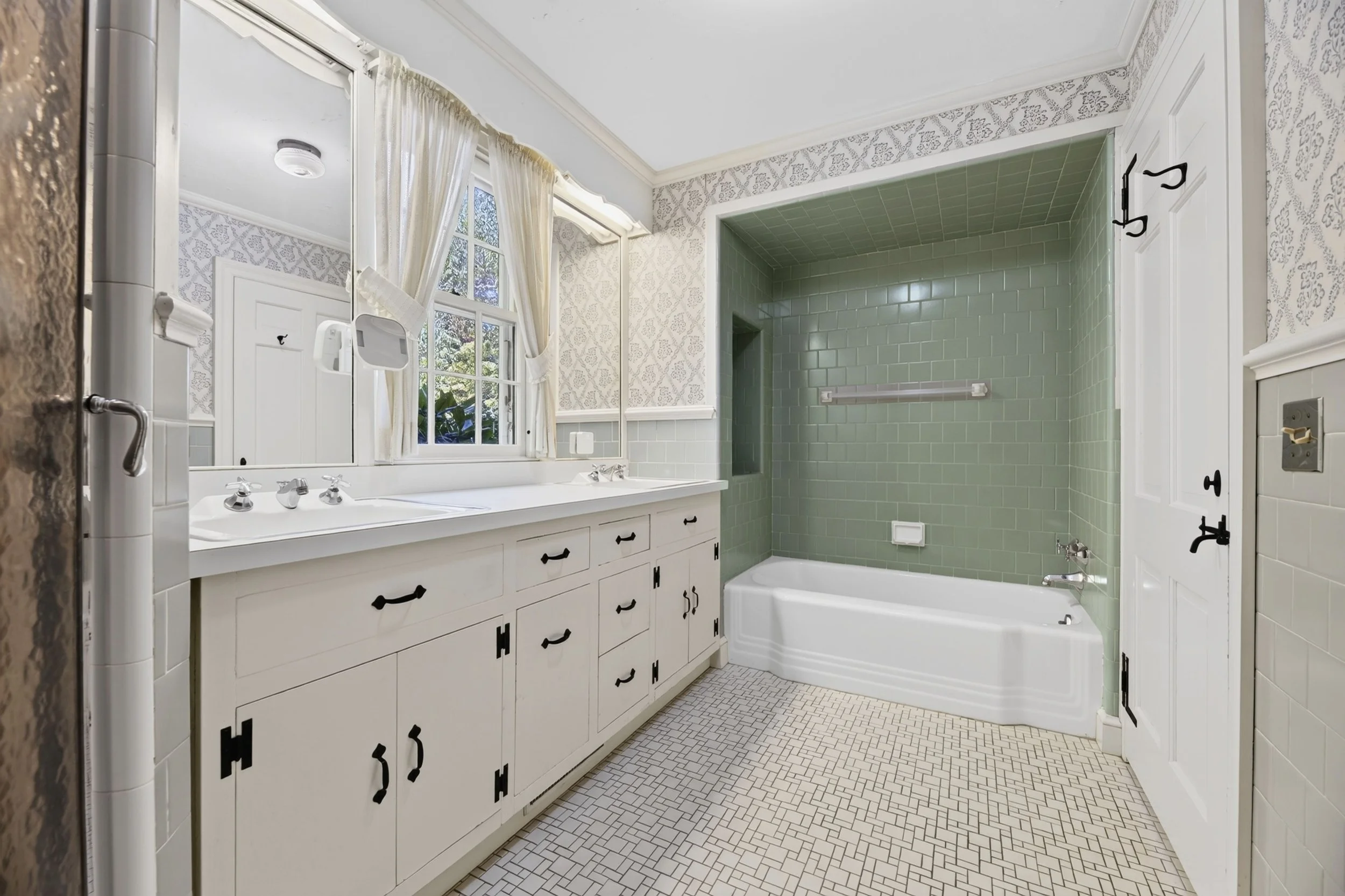 A vintage bathroom with a dual vanity, large mirror, and a green-tiled bathtub and shower area. Decor includes floral wallpaper, white curtains, and patterned tile flooring.