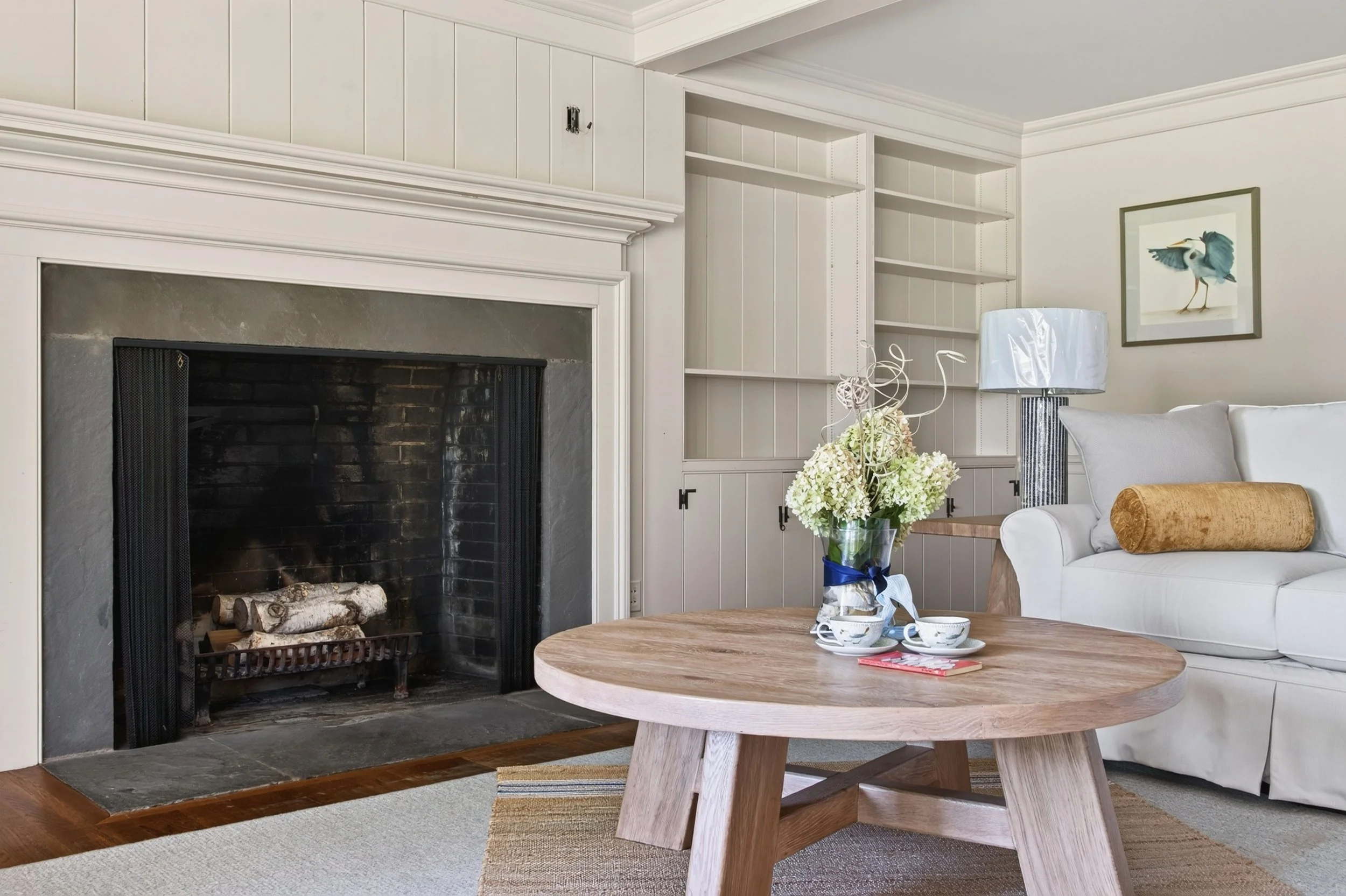 Living room with a white couch, coffee table, and a lamp. There is a fireplace with logs and a floral arrangement on the coffee table. Built-in shelves are empty, and a framed picture of a bird hangs on the wall.