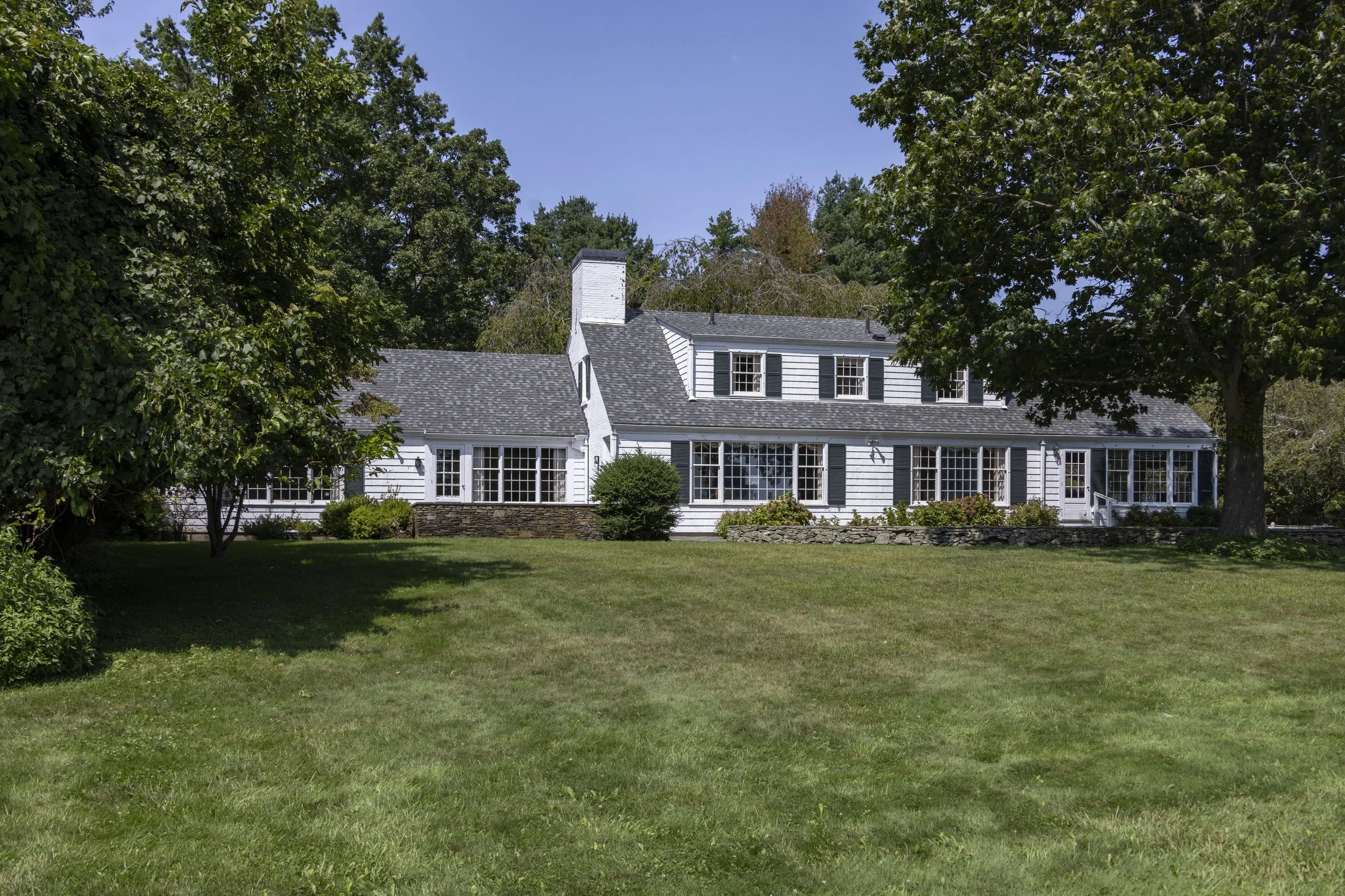 A white house with black shutters and a stone foundation, surrounded by green trees and a grassy yard, under a bright blue sky.