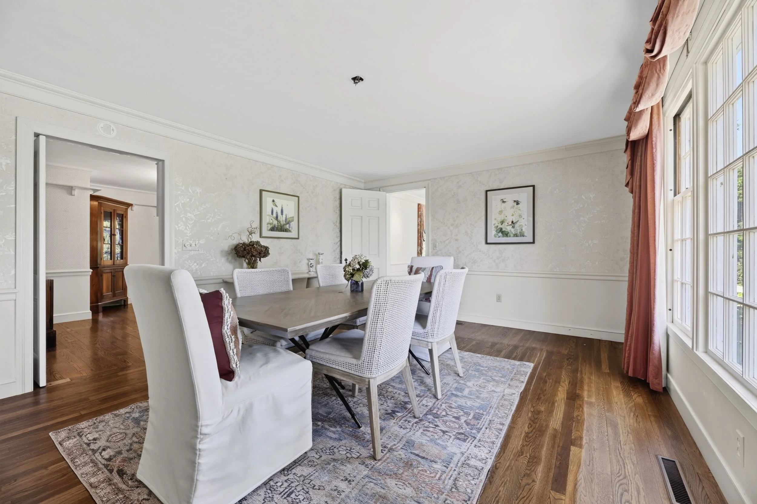A dining room with a wooden table surrounded by white upholstered chairs, a floral rug, framed botanical prints on the wall, a large window with pink curtains, and hardwood floors.