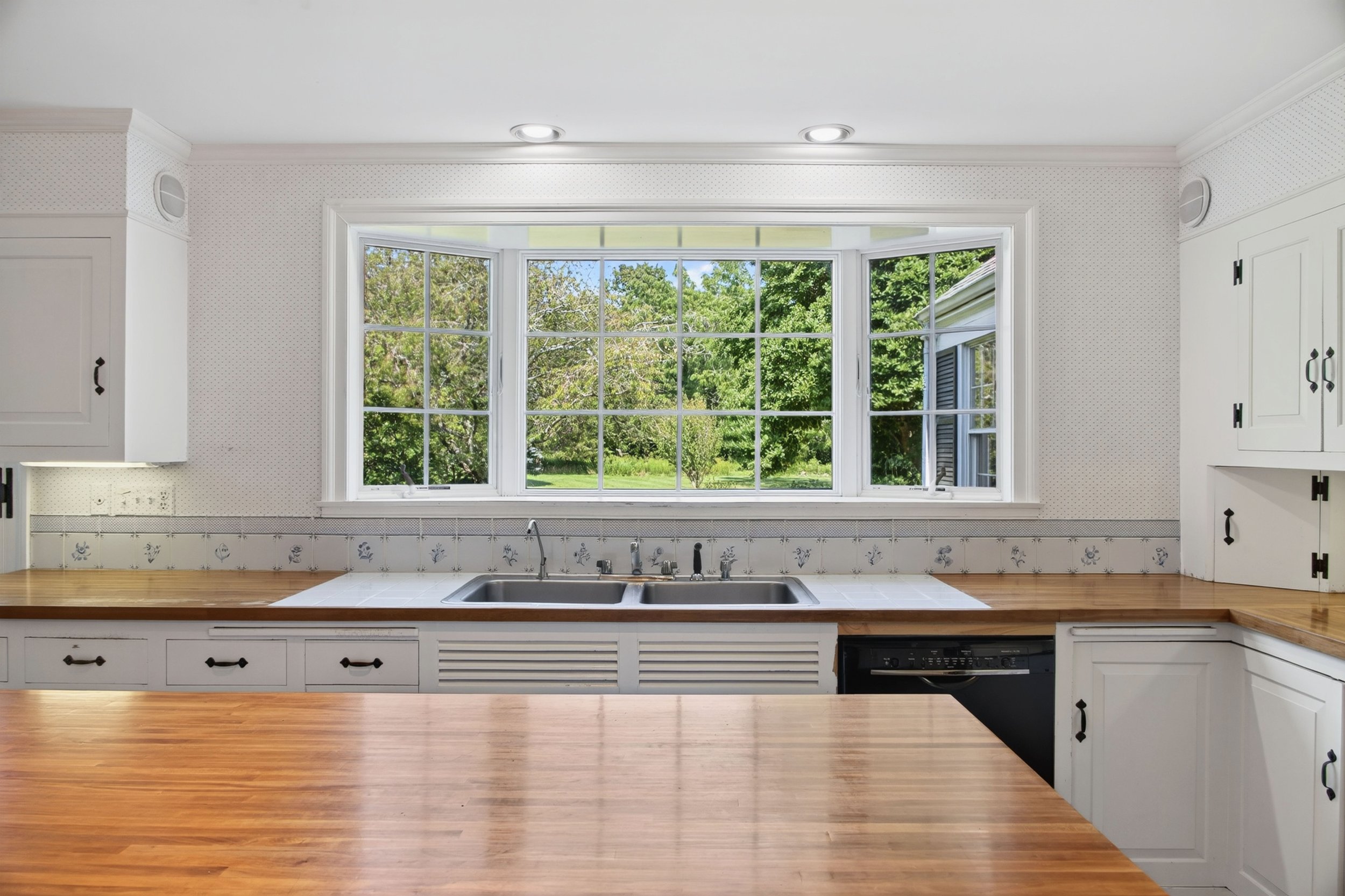 Kitchen with large window overlooking green trees and backyard, white cabinets with black hardware, wooden countertop, sink, and dishwasher.