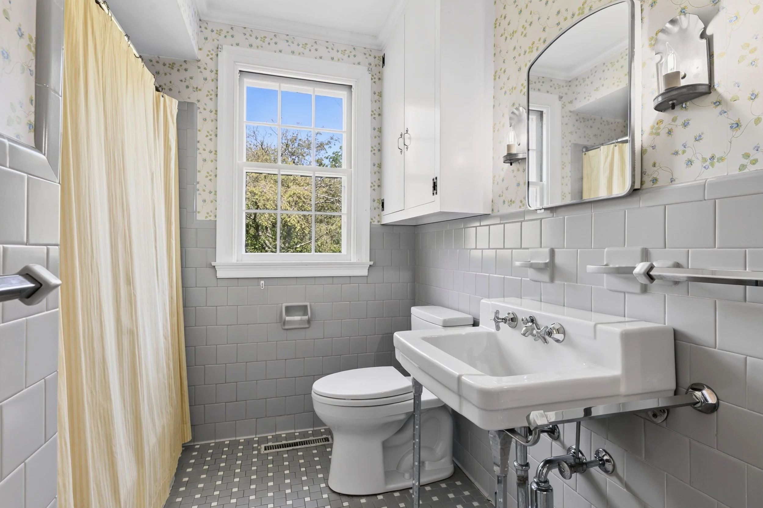 A small bathroom with white tiled walls, a large window showing trees outside, a white sink with vintage fixtures, a toilet, a mirror, and a yellow shower curtain.