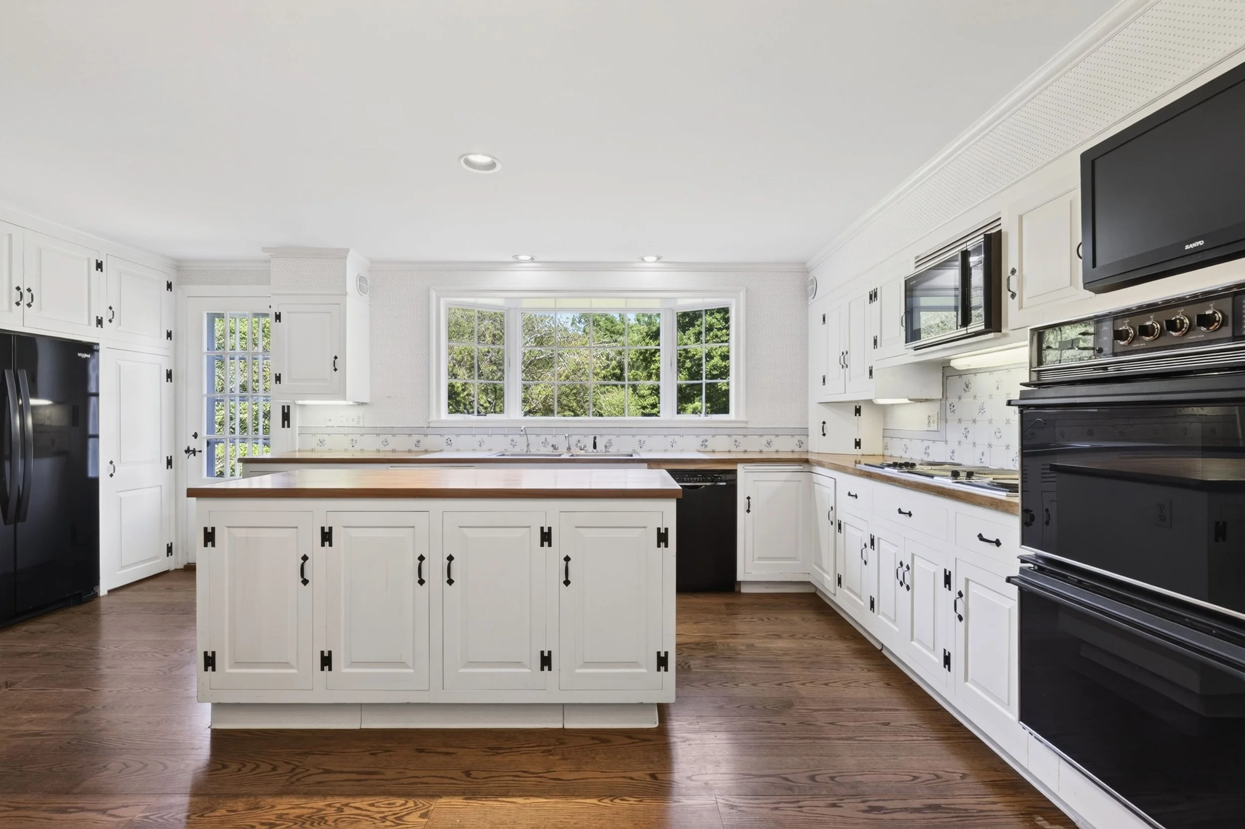 Bright white kitchen with large window, black appliances, and wood flooring.