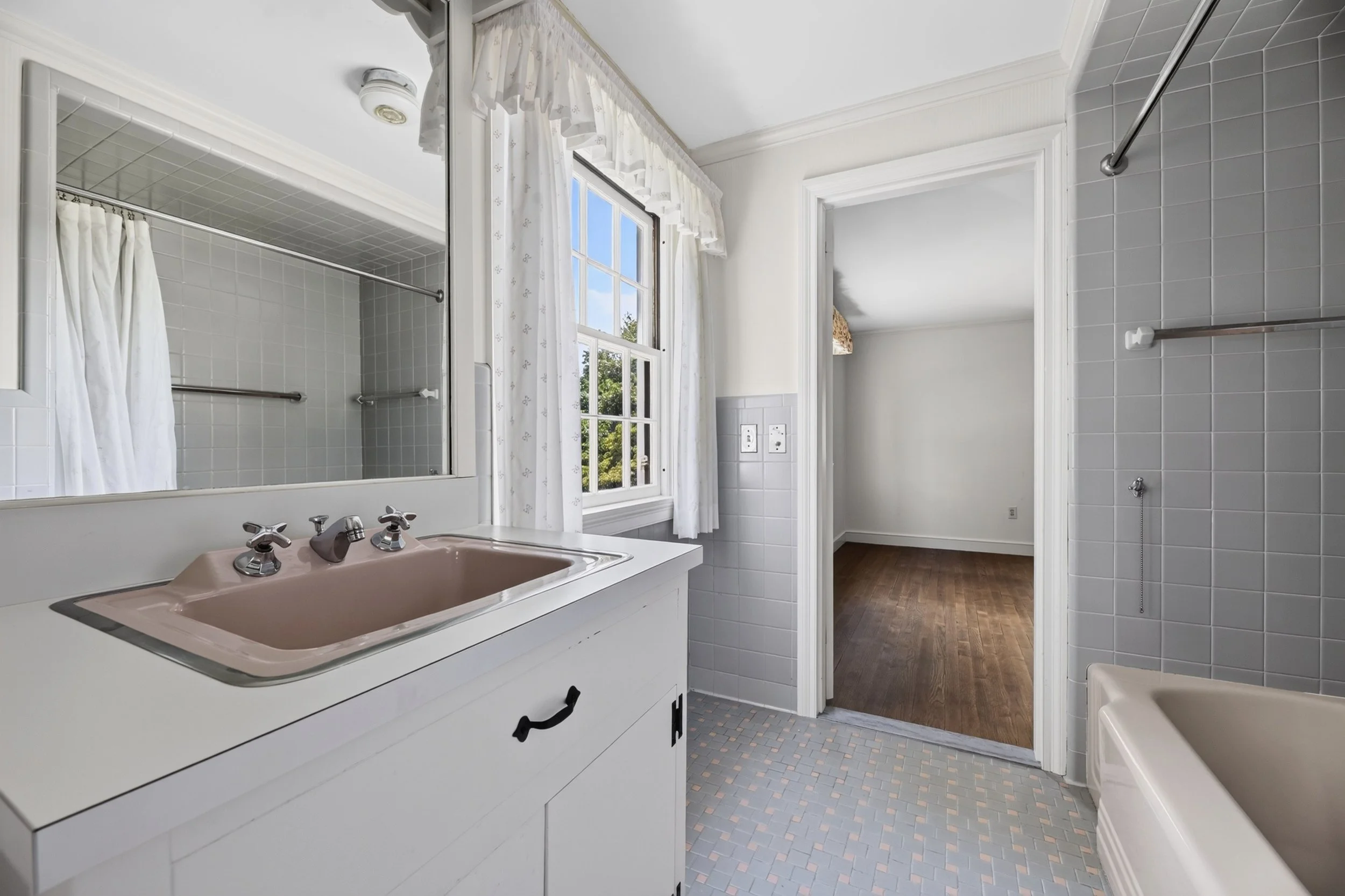 A bathroom with a pink sink, white cabinets, a mirror, a tiled shower area with a shower curtain, a window with white curtains, white walls, brown wooden flooring in another room visible through an open doorway, and tiled flooring in the bathroom.