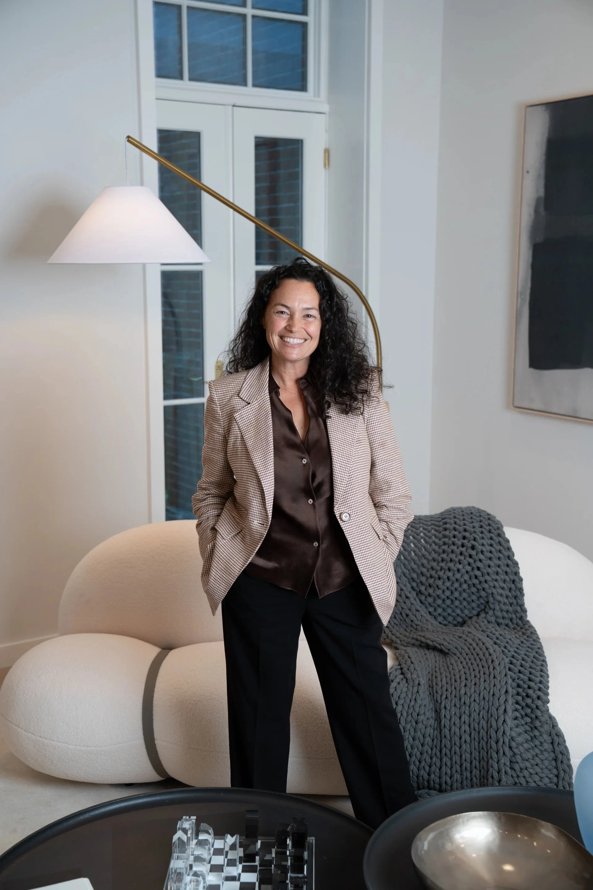 A woman with dark curly hair dressed in a checkered blazer and dark trousers standing in a modern living room next to a round beige sofa with a chunky gray throw blanket, smiling in front of a large window with white framing, a black and white modern art piece on the wall, and a black coffee table with glass decorative objects.