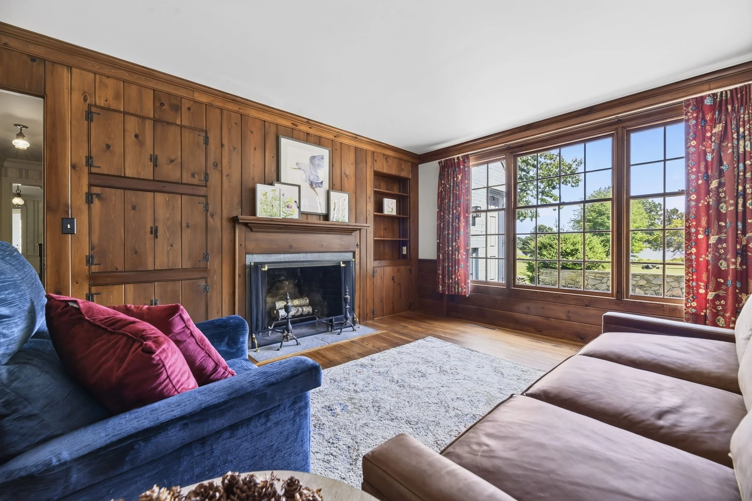 A living room with wood-paneled walls, a large window with floral curtains, a fireplace, and furniture including a blue velvet sofa with red pillows and a beige couch, sunlight streaming through the window.