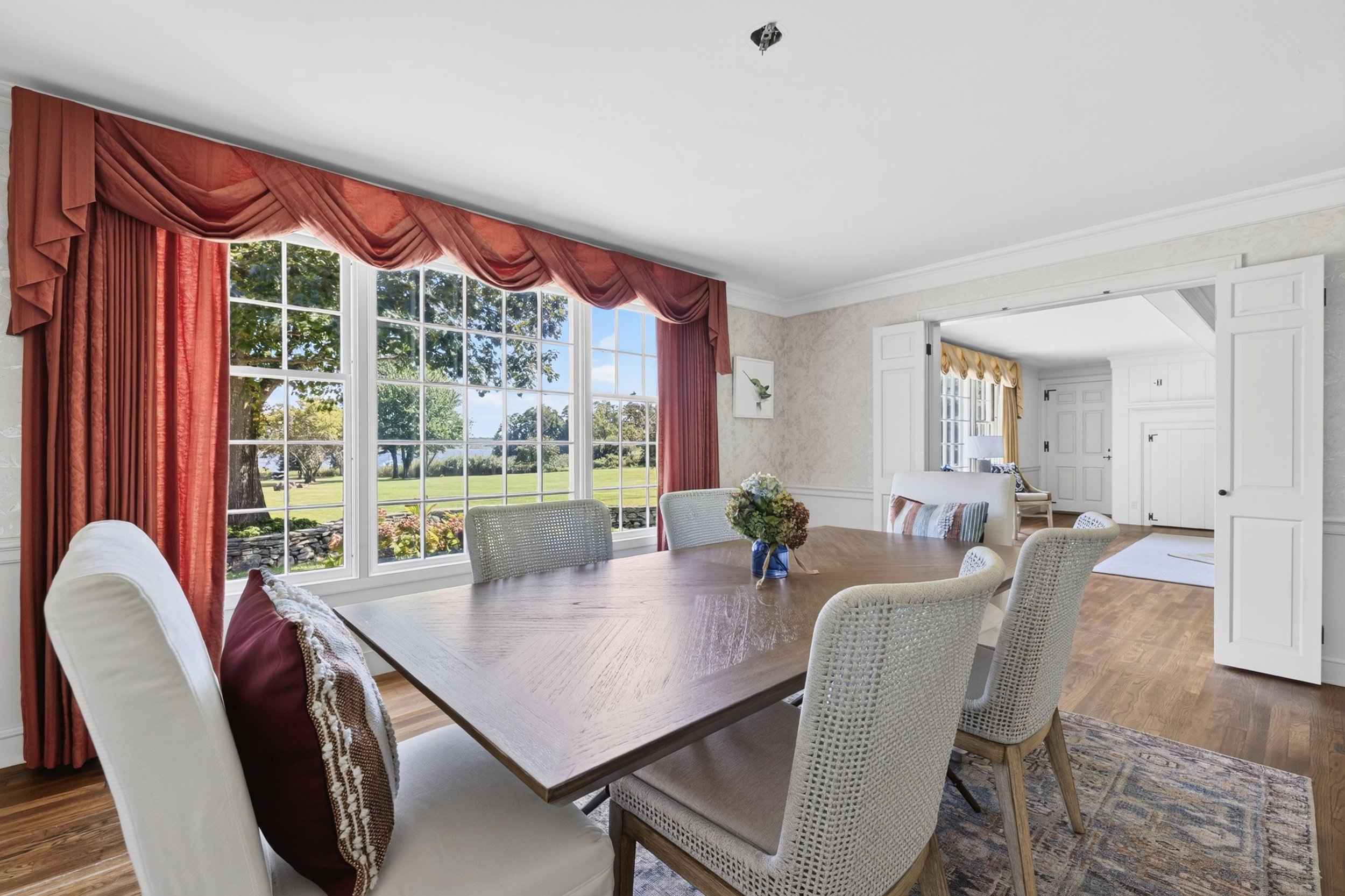 A dining room with large windows showing a green outdoor landscape, surrounded by red and yellow curtains, with a wooden dining table, white cushioned chairs, and a small flower arrangement in a blue vase.