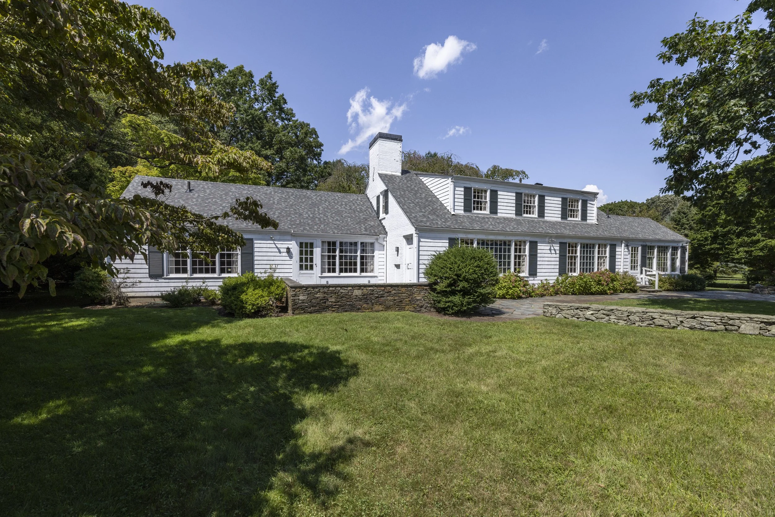 White house with black shutters, multiple windows, and a stone foundation, surrounded by greenery and trees under a blue sky with a few clouds.
