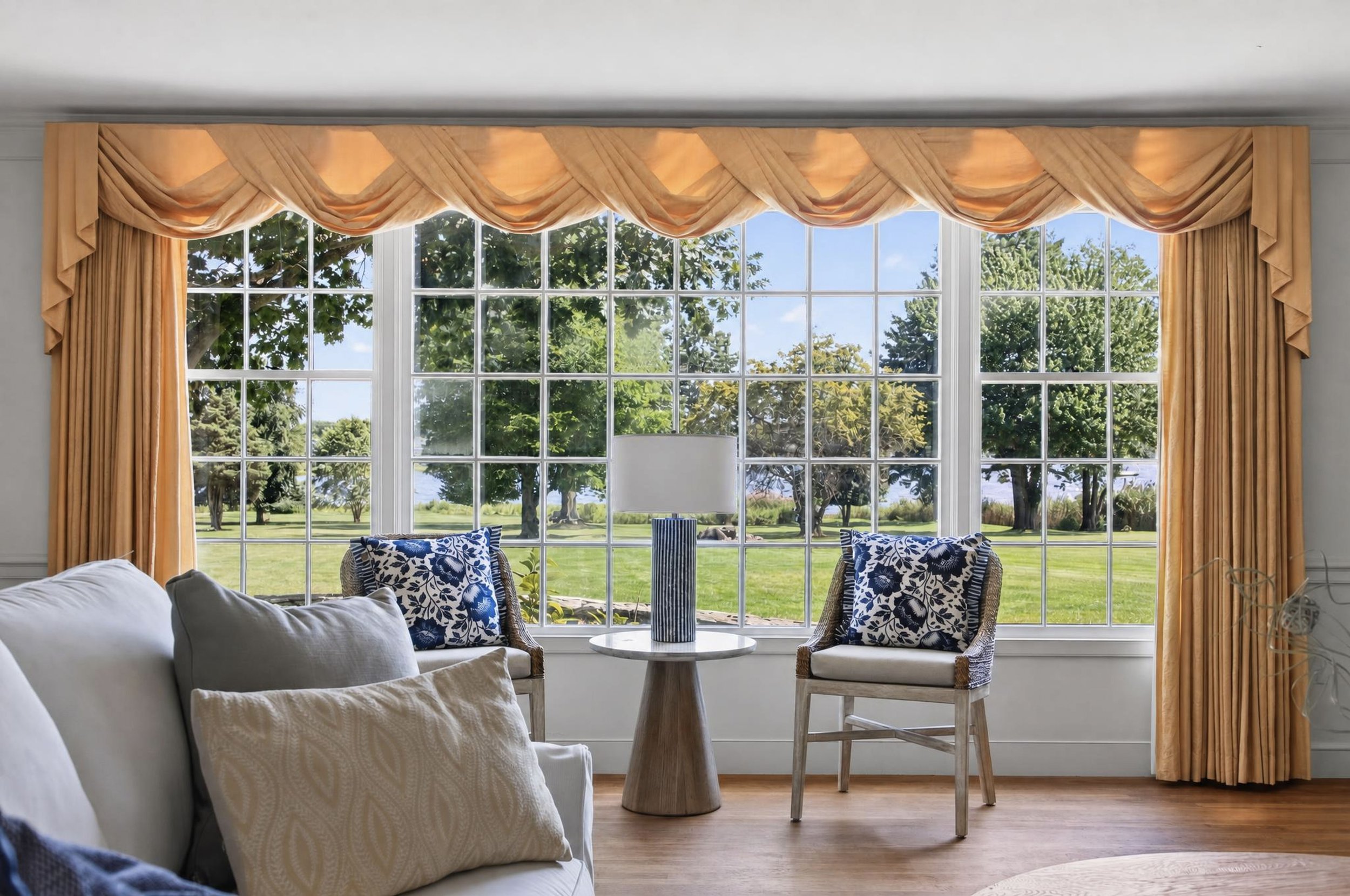 Living room with large window, peach curtains, white sofa with beige pillows, chairs with blue and white patterned cushions, and a small round table with a striped vase.