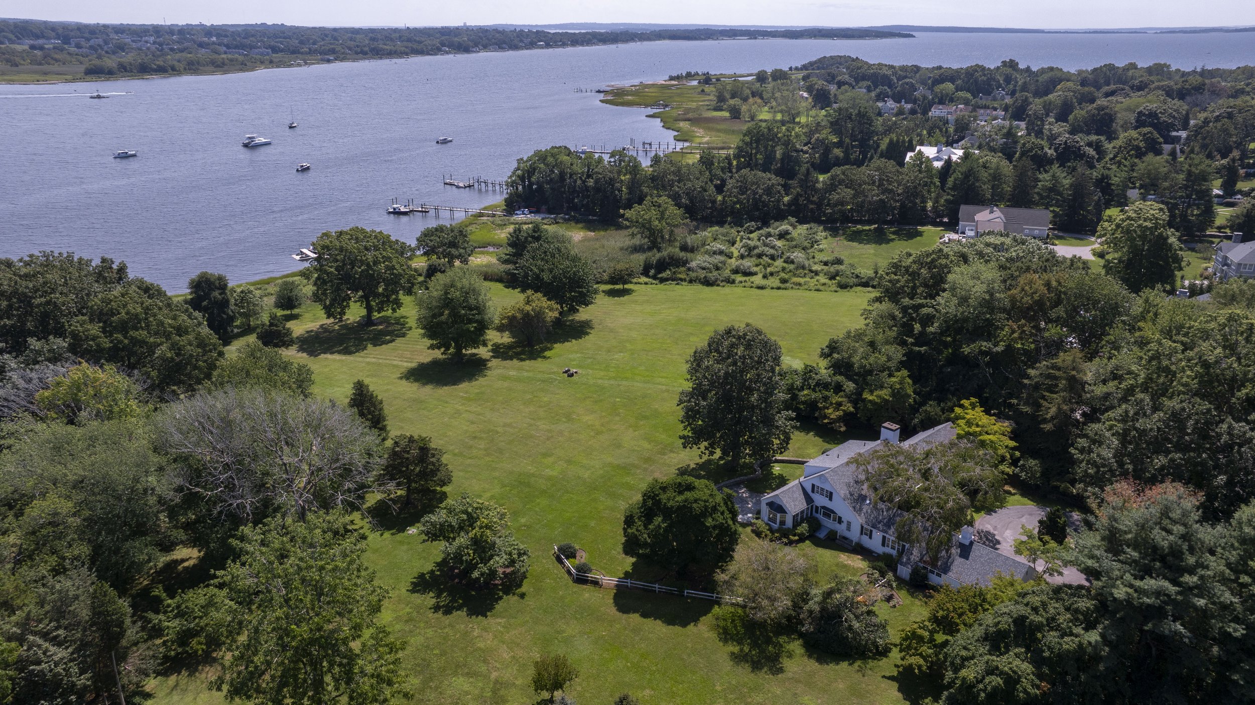 Aerial view of a waterfront with boats, green grassy land with trees, and residential houses along the shoreline.