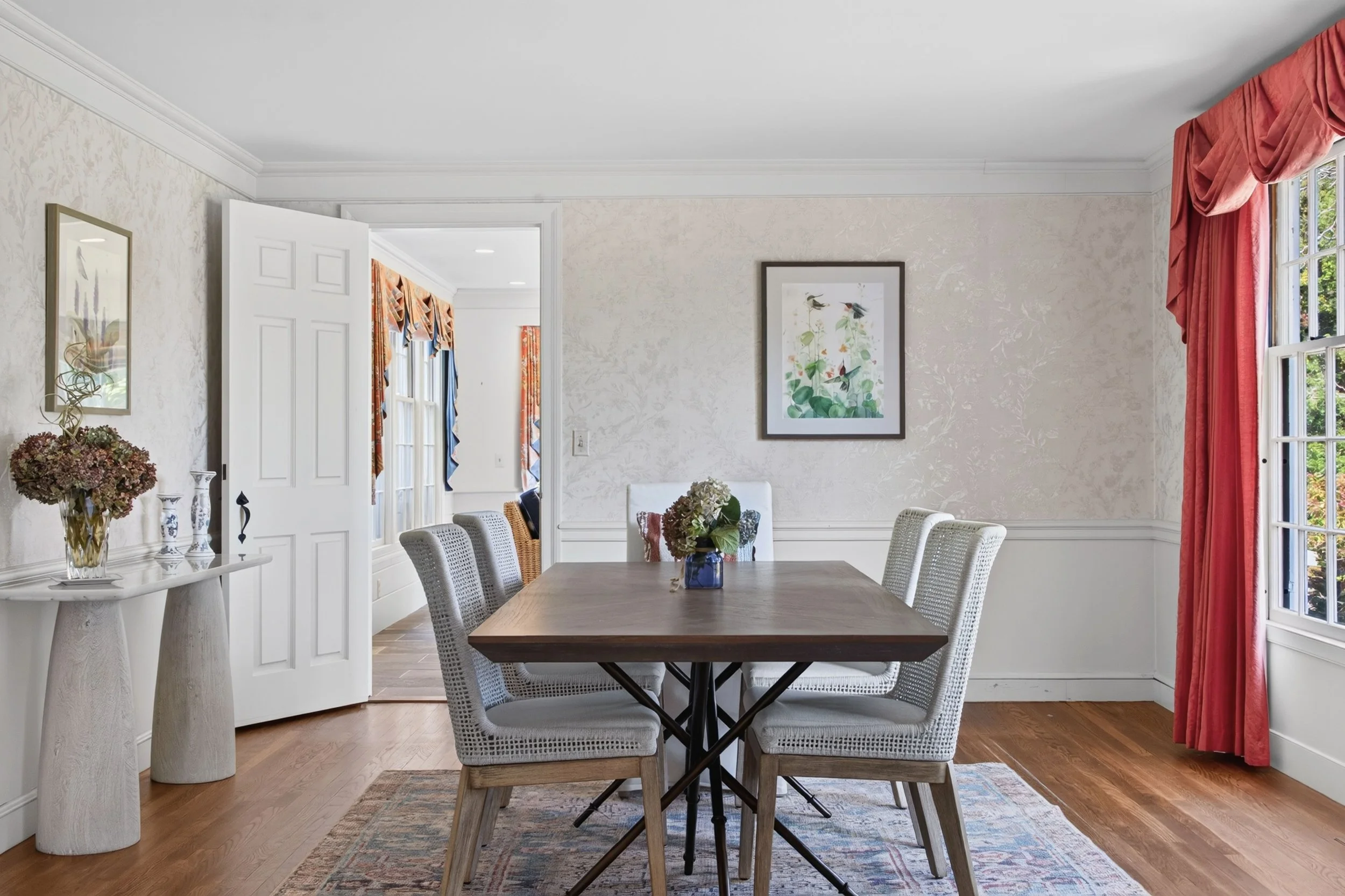 A dining room with a wooden table surrounded by white chairs, a window with pink curtains, framed bird artwork, a decorative floral arrangement, and a hallway with colorful curtains.