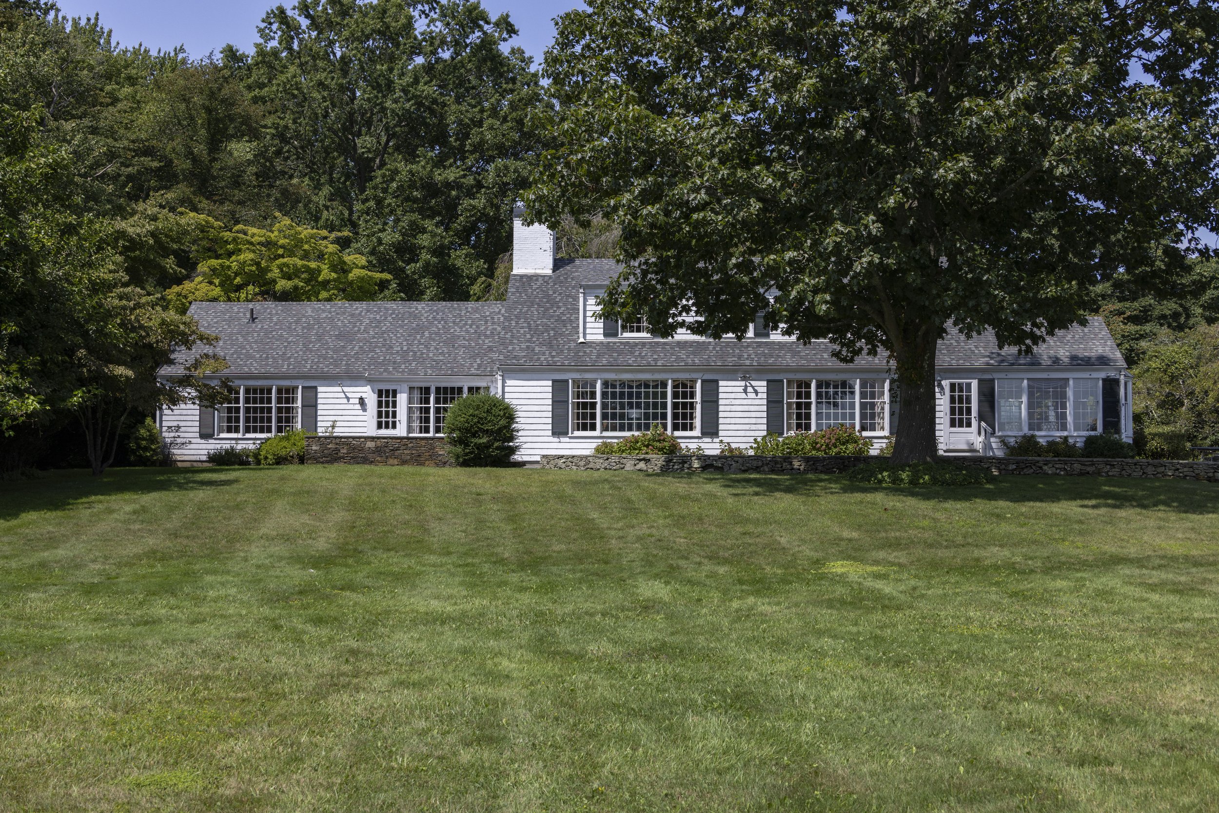 White house with multiple windows and shutters, surrounded by green trees and grass, with a clear blue sky in the background.