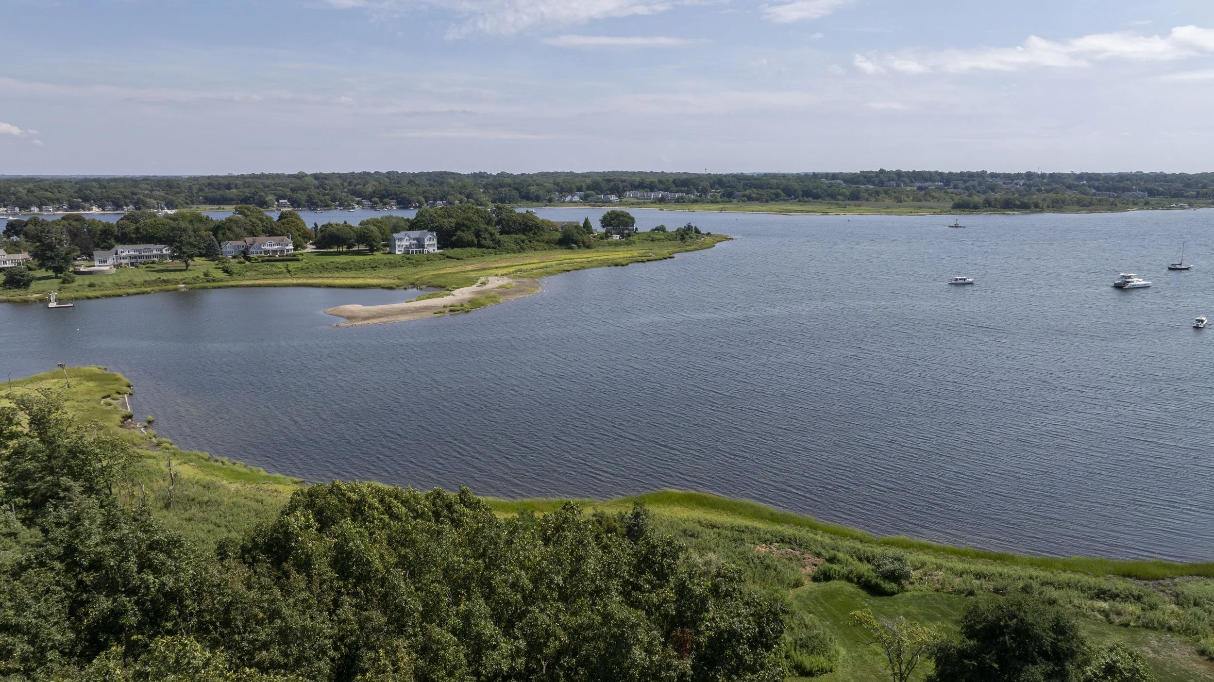 Aerial view of a coastal area with houses along the shoreline, boats on the water, and trees in the foreground under a partly cloudy sky.