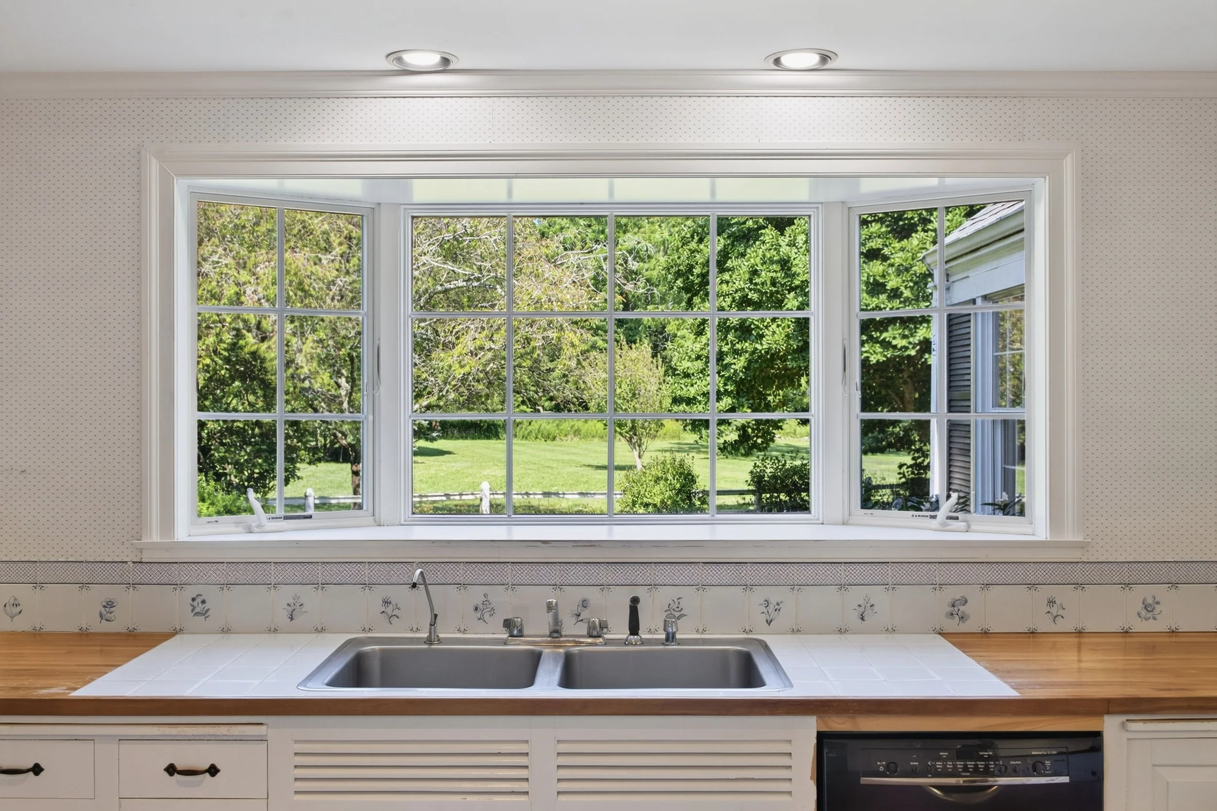 Kitchen view through a large window showing a green backyard with trees and grass, with a kitchen sink and countertop in the foreground.