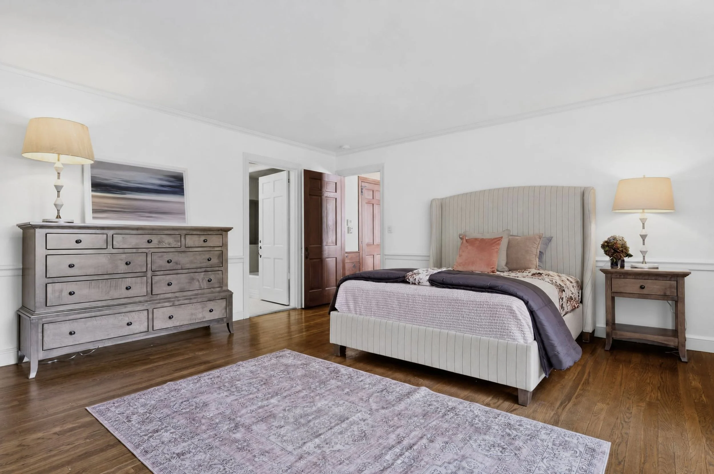 A bedroom with a bed, two nightstands with lamps, a dresser, a framed picture, and wooden floors.