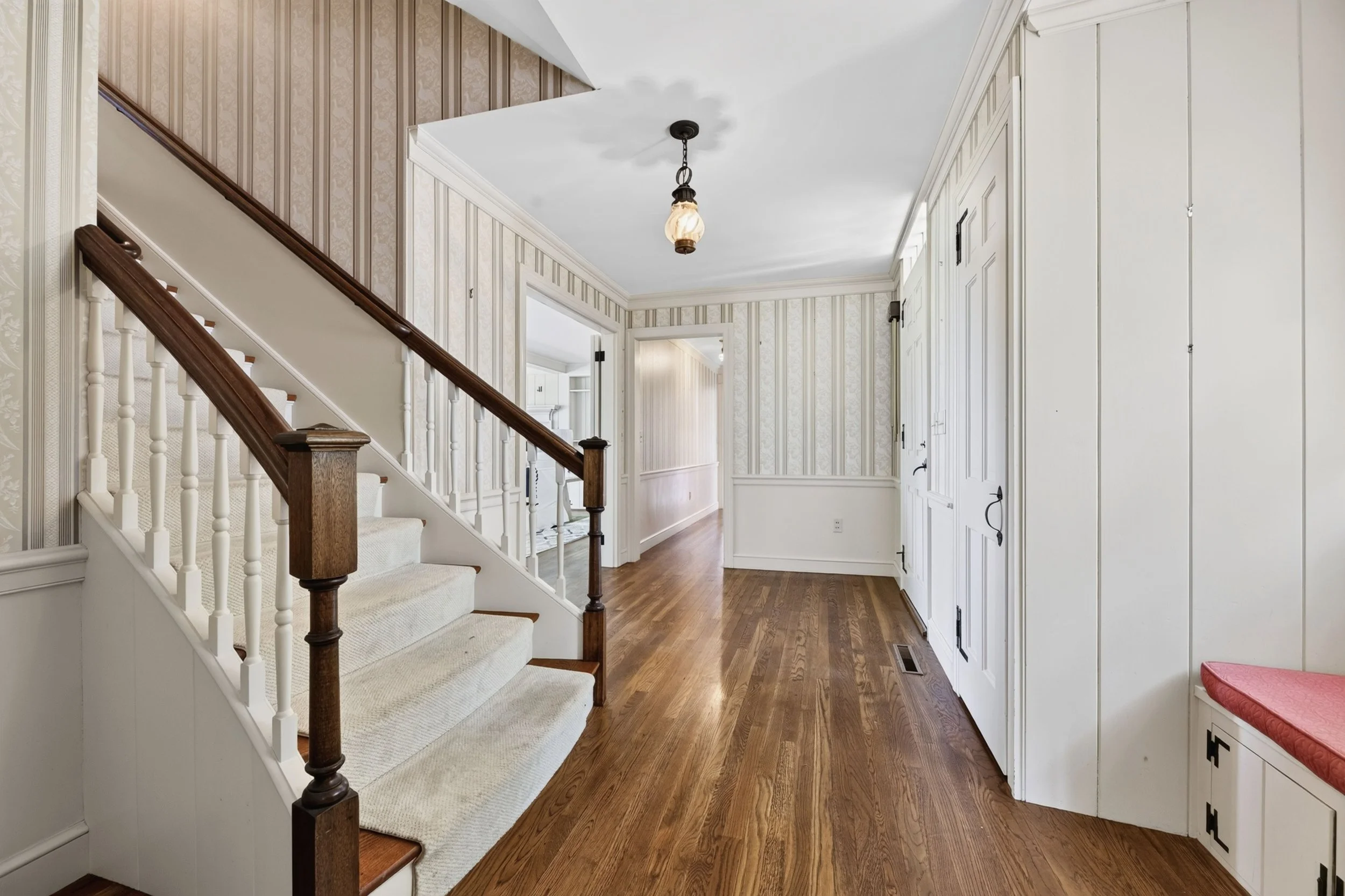 Interior view of a home entrance with staircase on the left, hardwood flooring, floral patterned wallpaper, a ceiling hanging light, and white cabinets on the right.