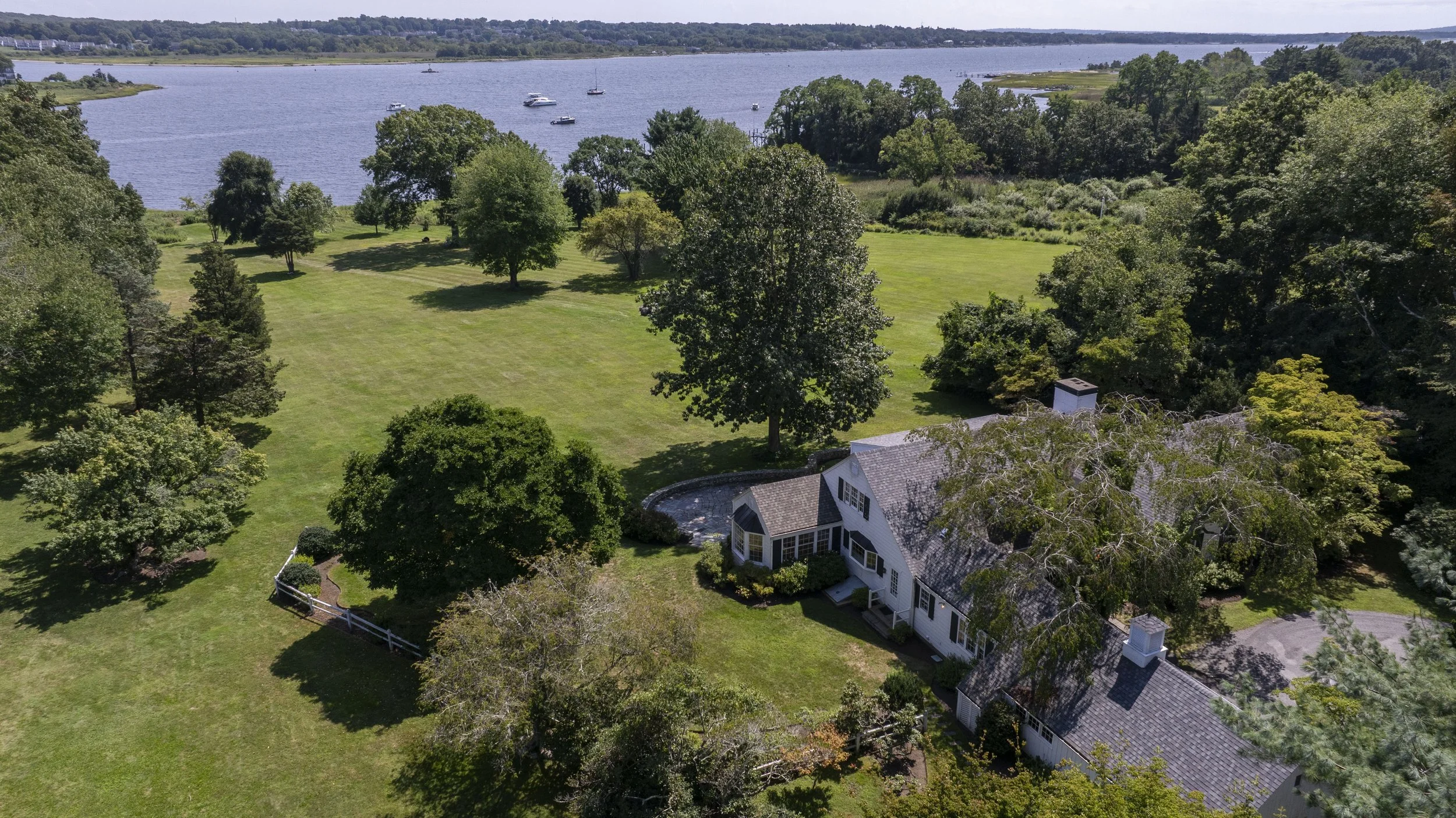 Aerial view of a large house with a well-maintained lawn, surrounded by trees, with a river in the background and boats floating on it.