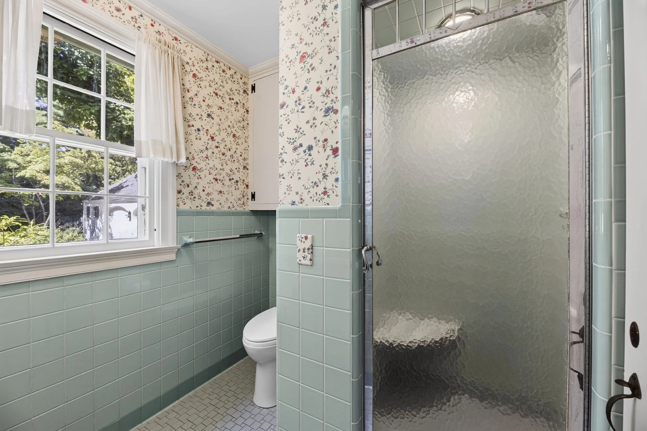 A vintage-style bathroom with mint green tiled walls, a toilet, a glass shower stall, a window with white curtains, and floral wallpaper.