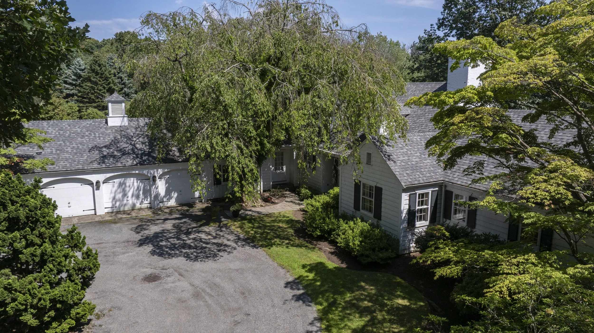 Aerial view of a white house with black shutters, a gray shingled roof, surrounded by green trees and shrubbery, with a driveway in front.