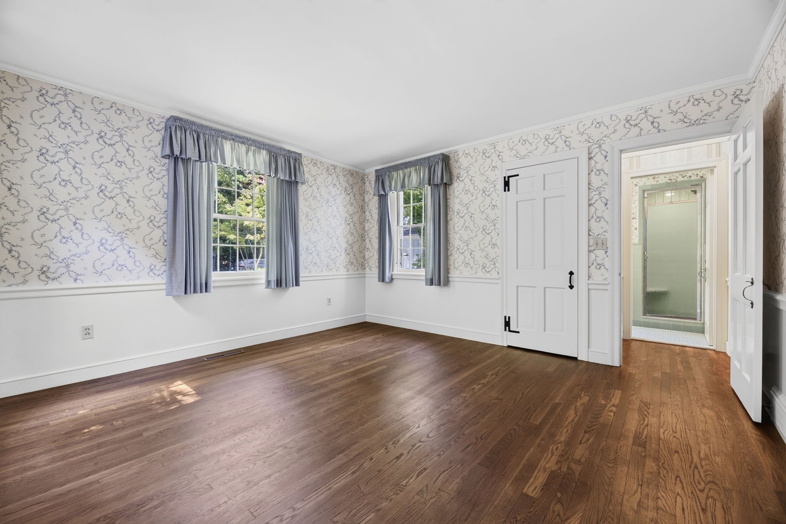 An empty room with hardwood floors, wallpapered walls with a light floral pattern, two windows with blue curtains and valances, a white door, and a view into a bathroom with teal tile and a frosted glass shower door.