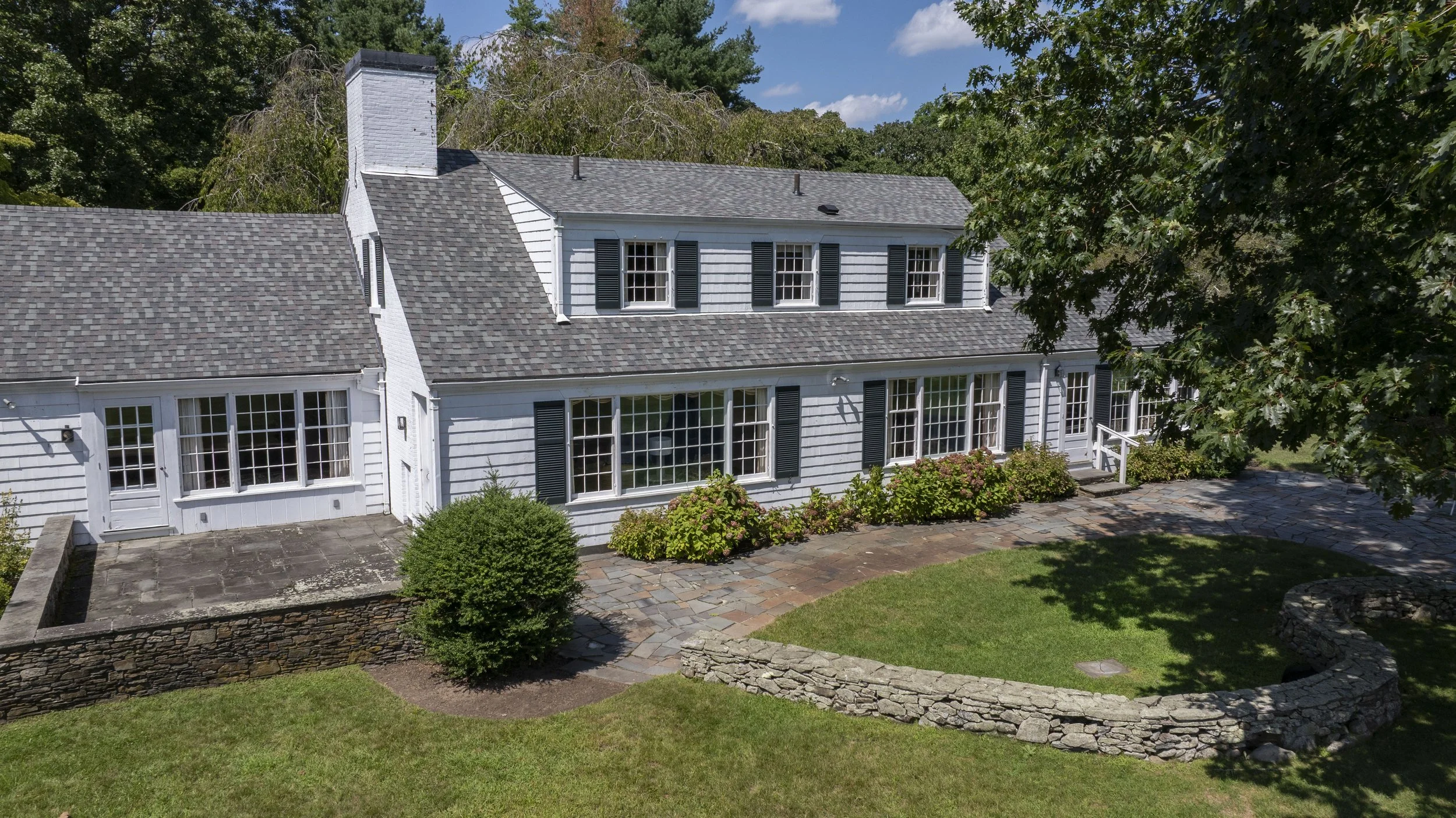 A large two-story white house with black shutters and a slate roof, surrounded by green trees and a landscaped backyard with a stone patio and a small retaining wall.