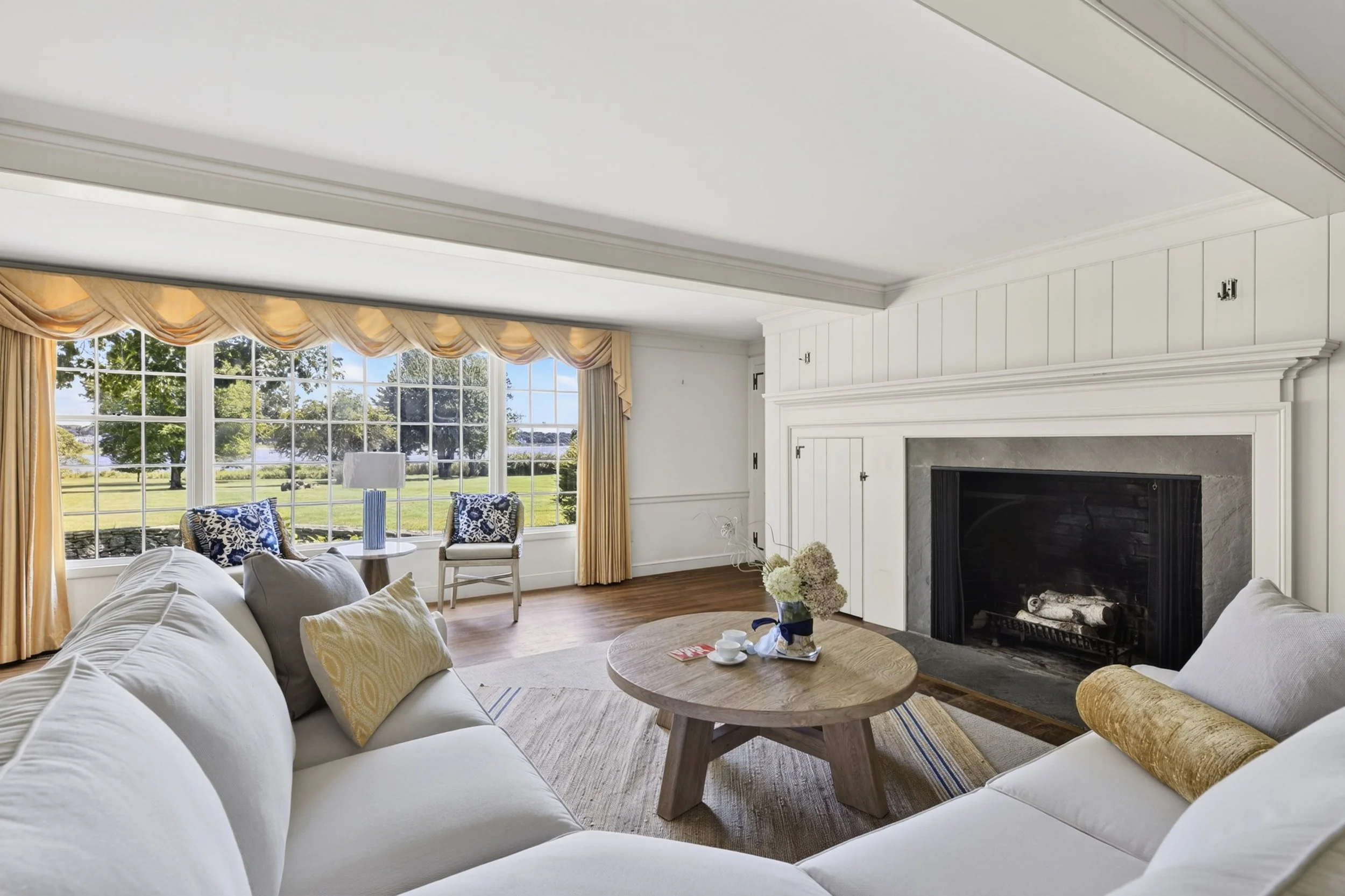 A living room with a white sofa and colorful pillows, wooden coffee table, large window with beige curtains showing a lake and trees outside, a fireplace with a white mantel, and floral arrangement on the floor.