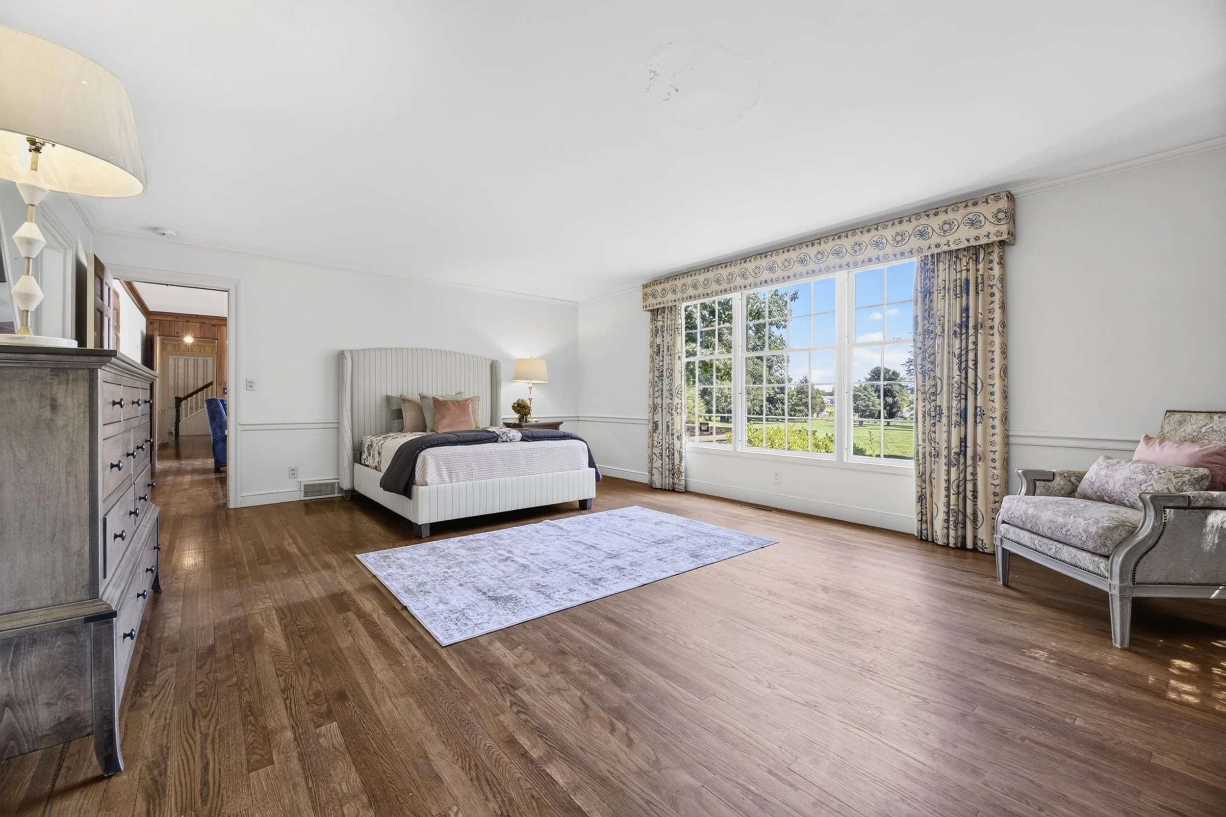 A spacious bedroom with hardwood floors, a white bed with pillows, a rug, a dresser with a lamp, large windows with patterned curtains, and a view of trees and a grassy area outside.