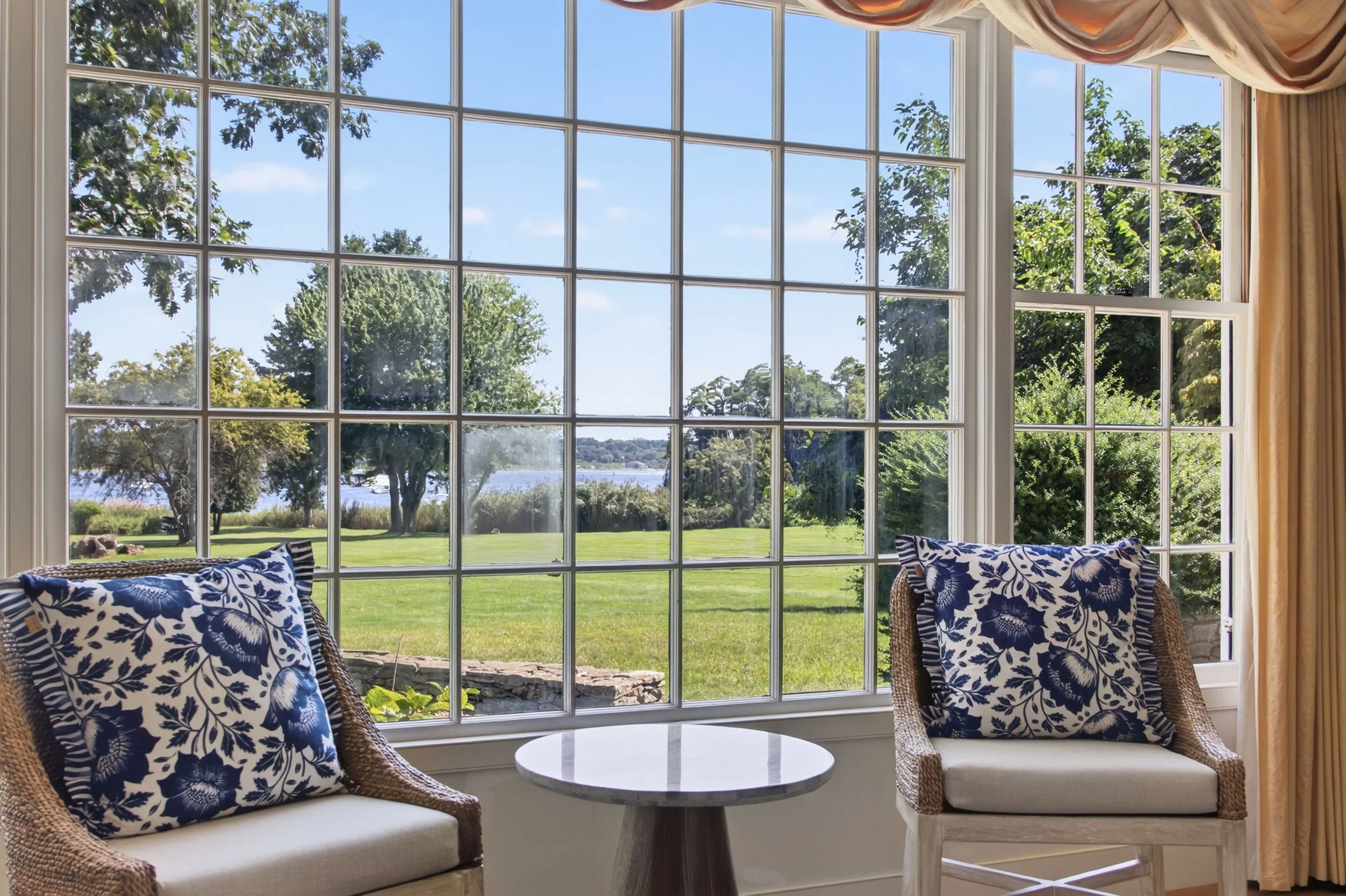 Room with large window overlooking a green lawn and trees, with curtains pushed to the side. Two wicker chairs with blue floral cushions and a small white round table are visible.