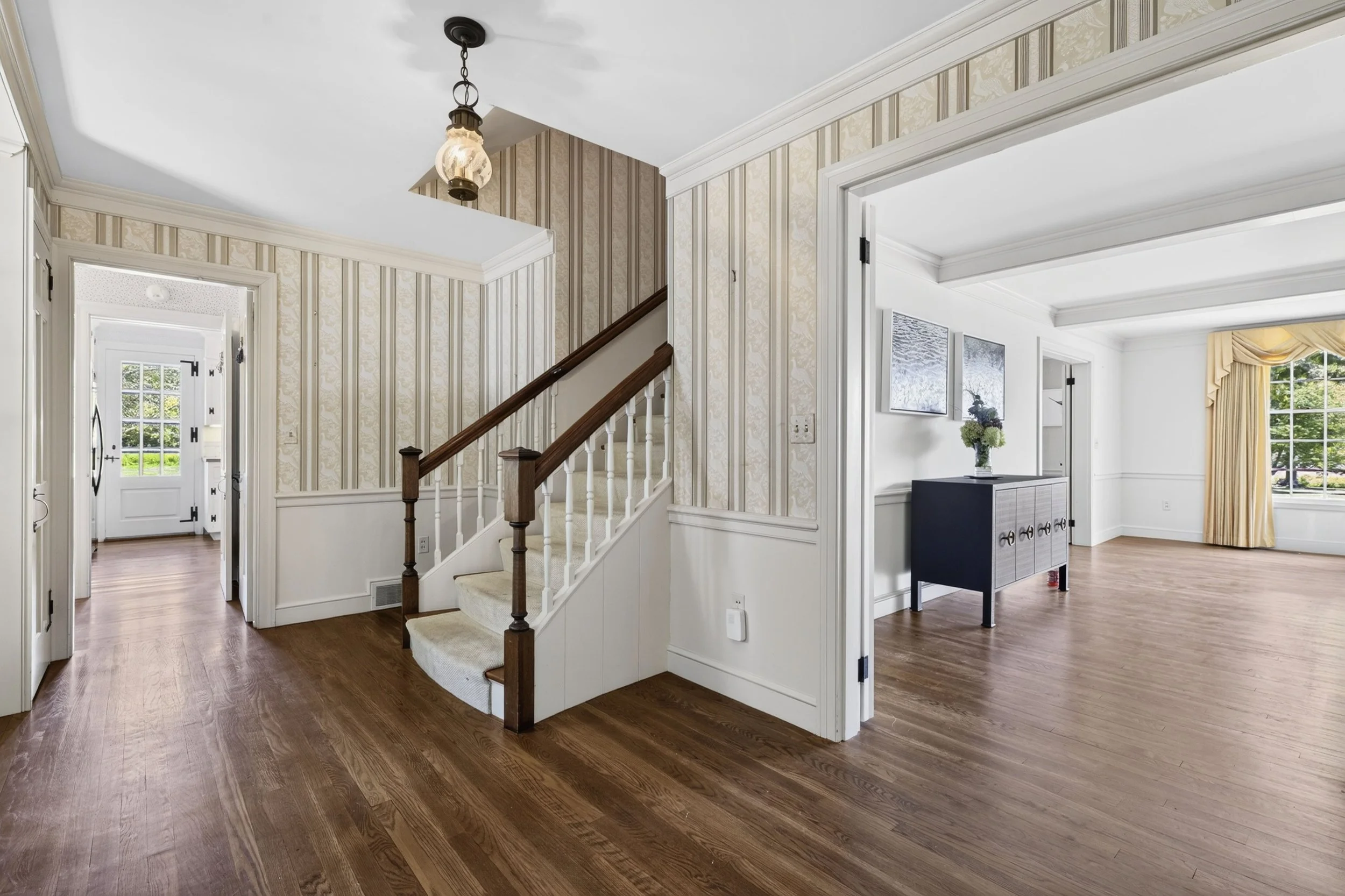 Interior view of a home with hardwood floors, a staircase with a wooden handrail, a doorway leading to a room with a black cabinet, and a large window with yellow curtains.