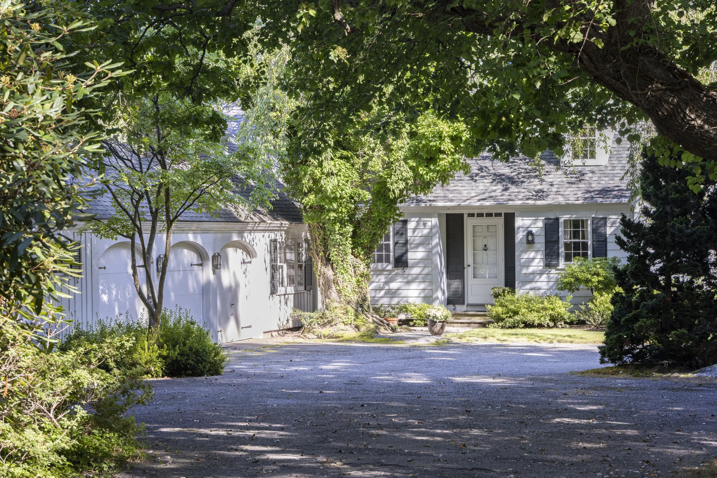 A white house with black shutters and a gray shingle roof, partially obscured by large green trees and bushes, with a driveway leading to a garage door and a front door, in a sunny yard.