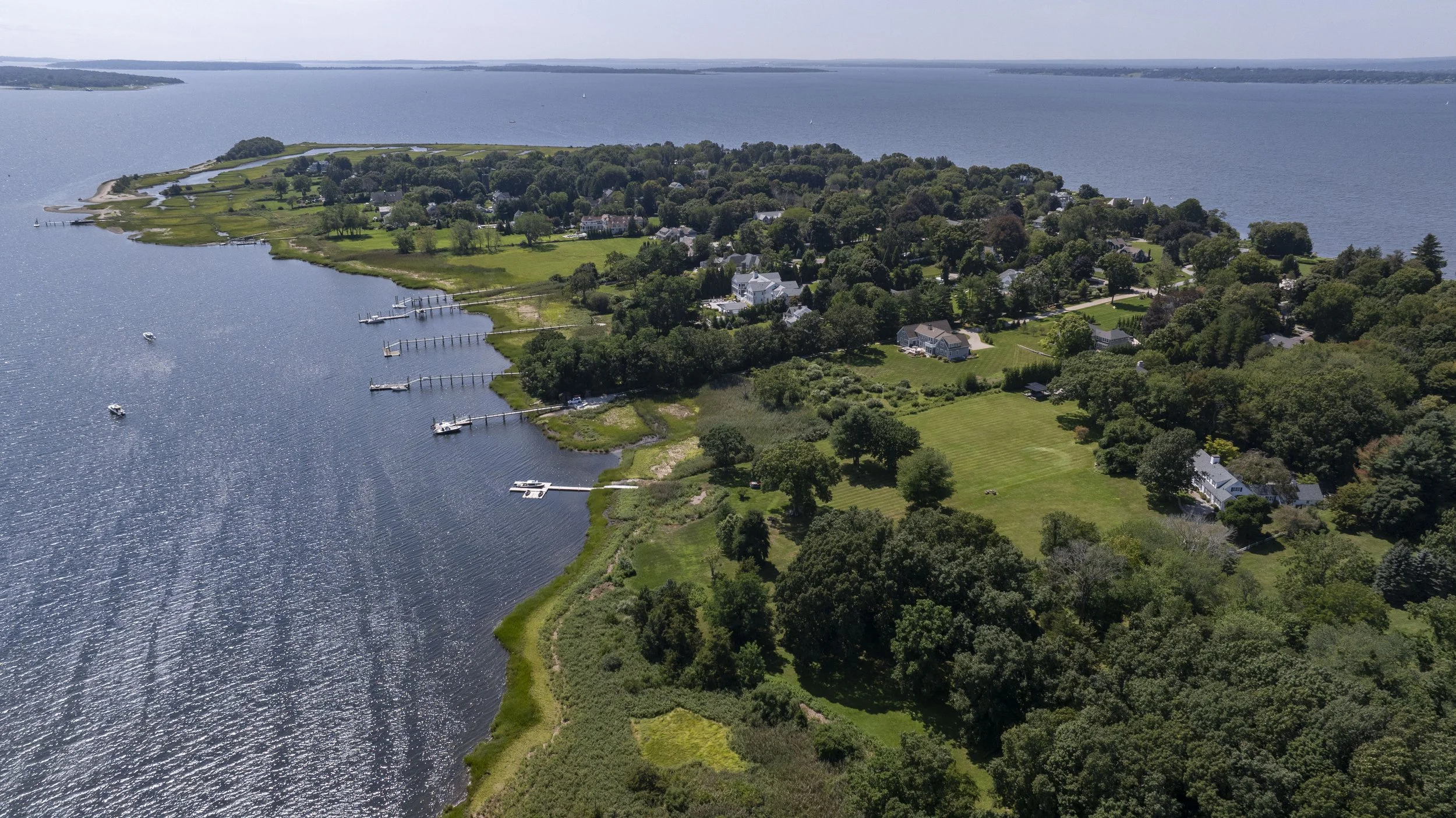 Aerial view of a coastal residential area with houses, docks, and boats along a large body of water, surrounded by greenery and trees.