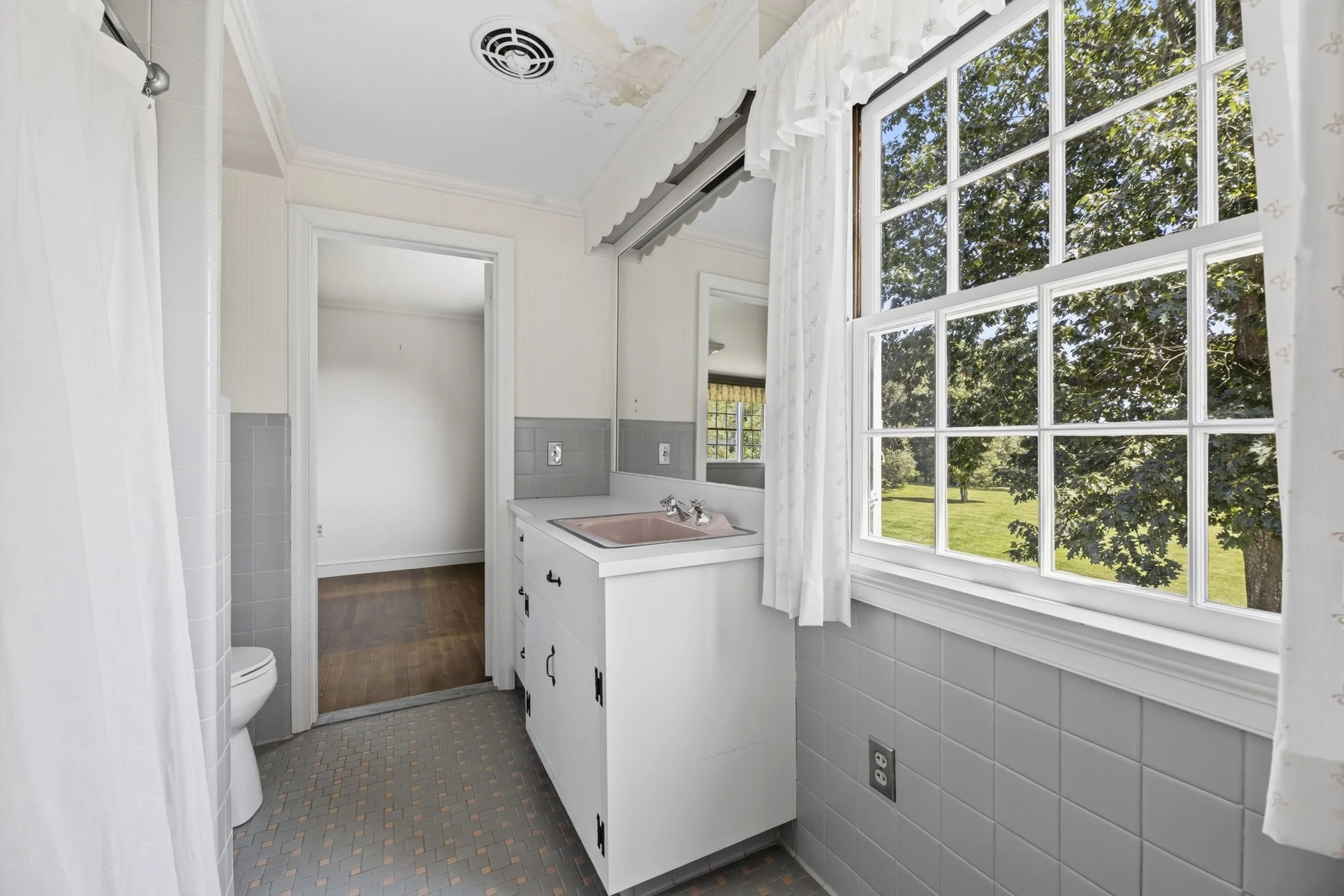 Old bathroom with peeling ceiling paint, large window with white curtains, pink sink, white cabinet, and gray tile floor.