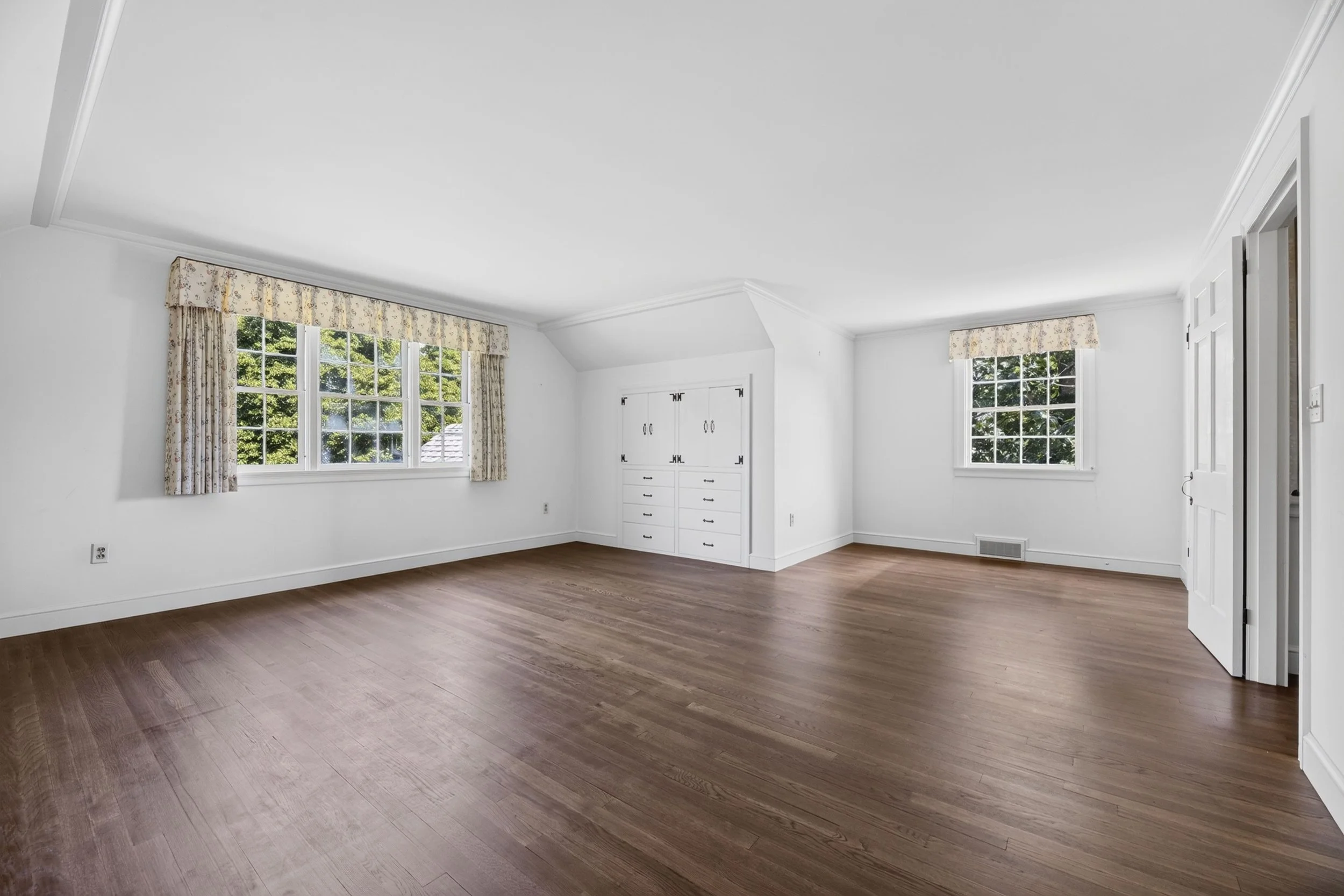Empty room with white walls, hardwood floors, large windows with floral curtains, and built-in white storage cabinet.
