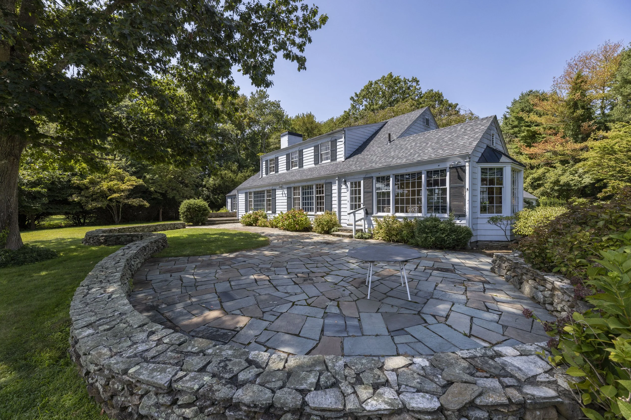 White house with black shutters and a large porch, surrounded by trees and greenery, with a stone patio in the foreground and a clear blue sky above.