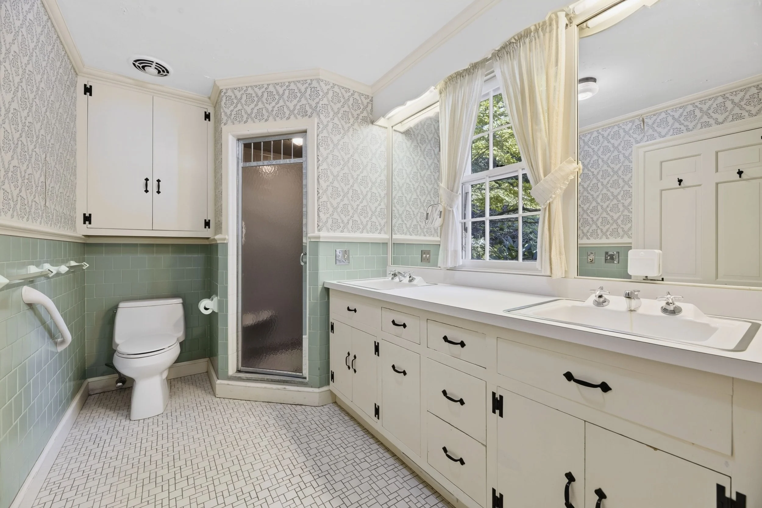 A vintage bathroom with white cabinets, a double sink, a window with cream curtains, a toilet with grab bars, a frosted glass shower door, and green tile on the walls.