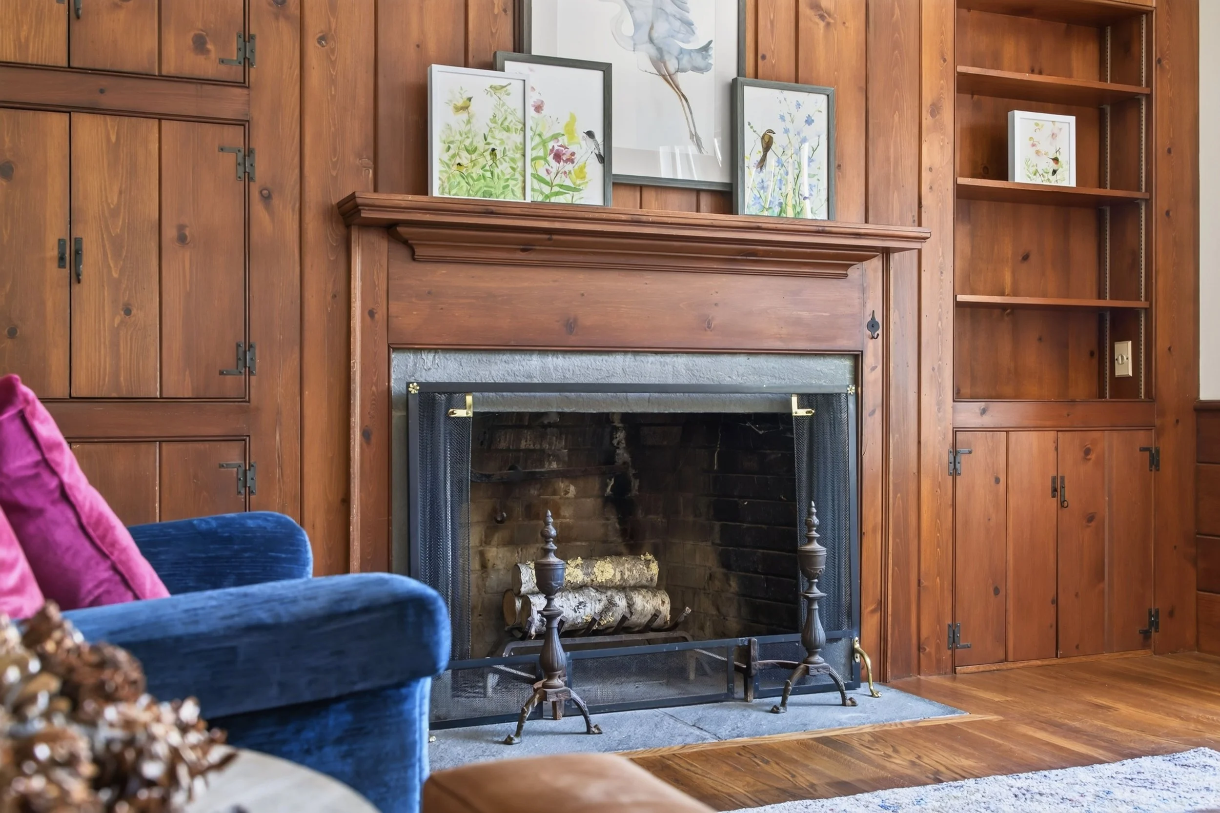 Living room with wood-paneled walls, a fireplace with logs, and framed bird watercolor paintings above the mantel. Blue velvet sofa with pink and purple cushions is partially visible in the foreground.