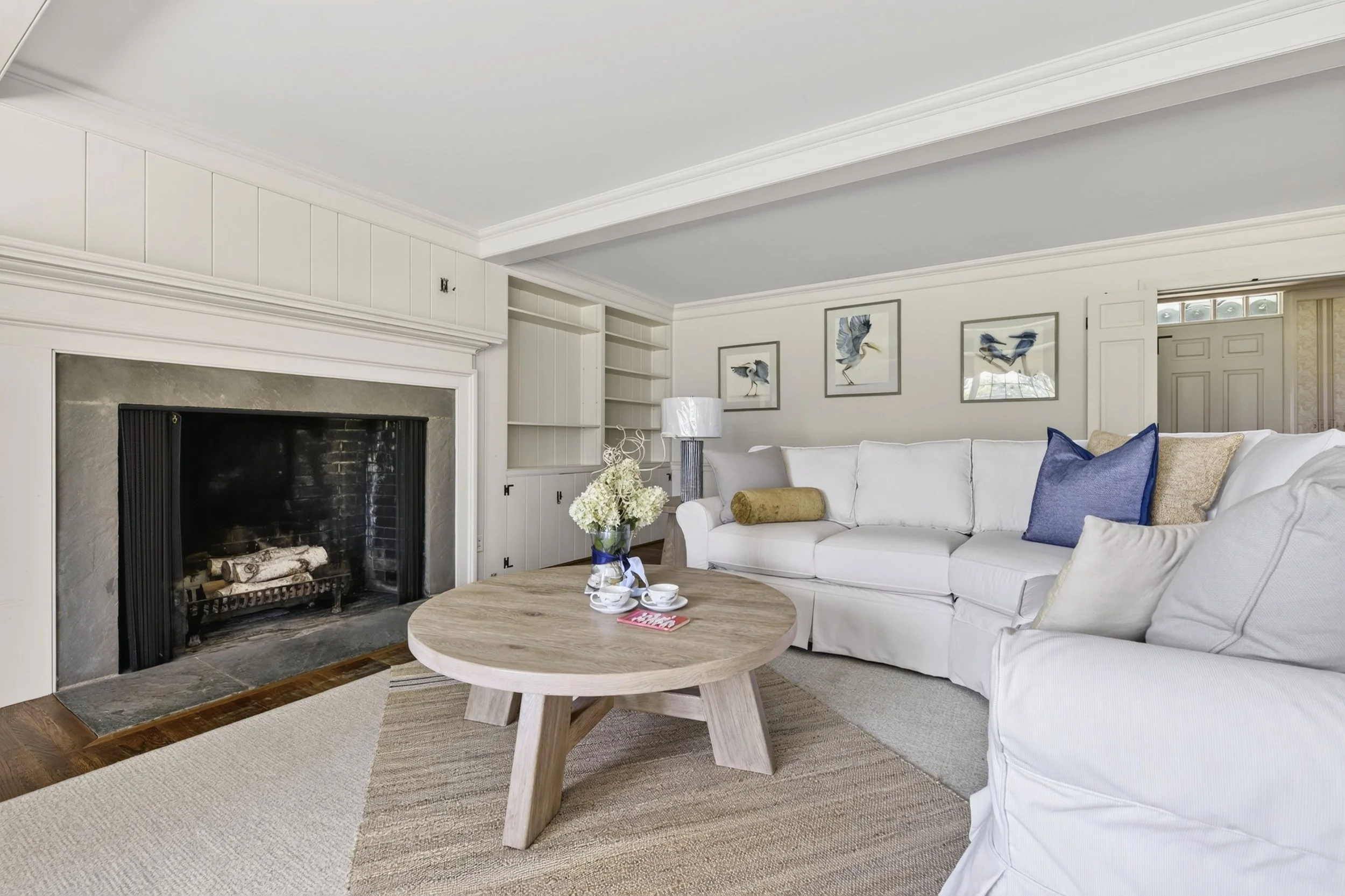 A living room with a white sofa, a wooden coffee table with a vase of white flowers, a fireplace, built-in shelves, framed bird artwork on the walls, a lamp, and a window with shutters.