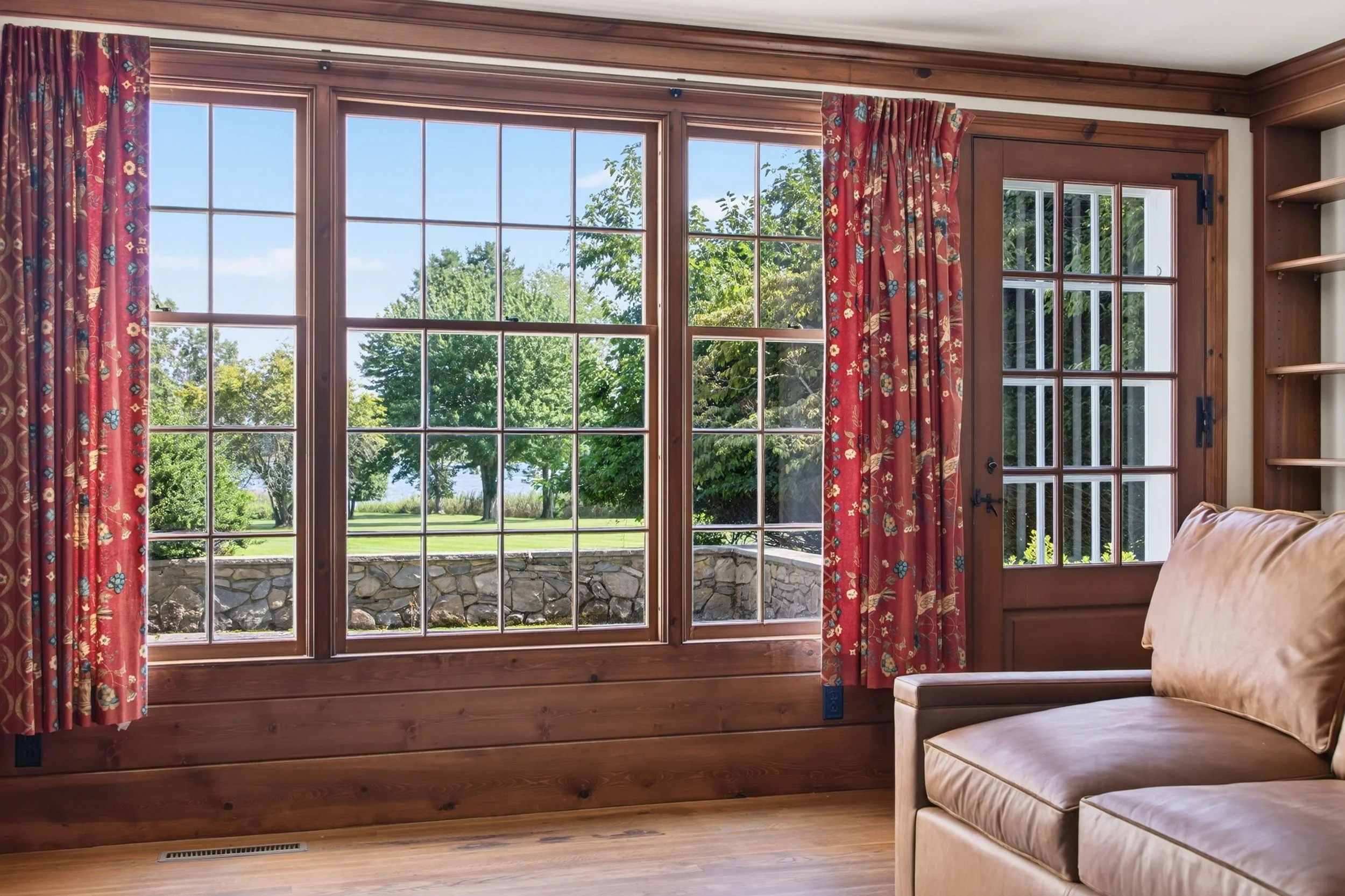 Wooden framed windows with red floral curtains looking out onto a green yard with trees and a stone wall.