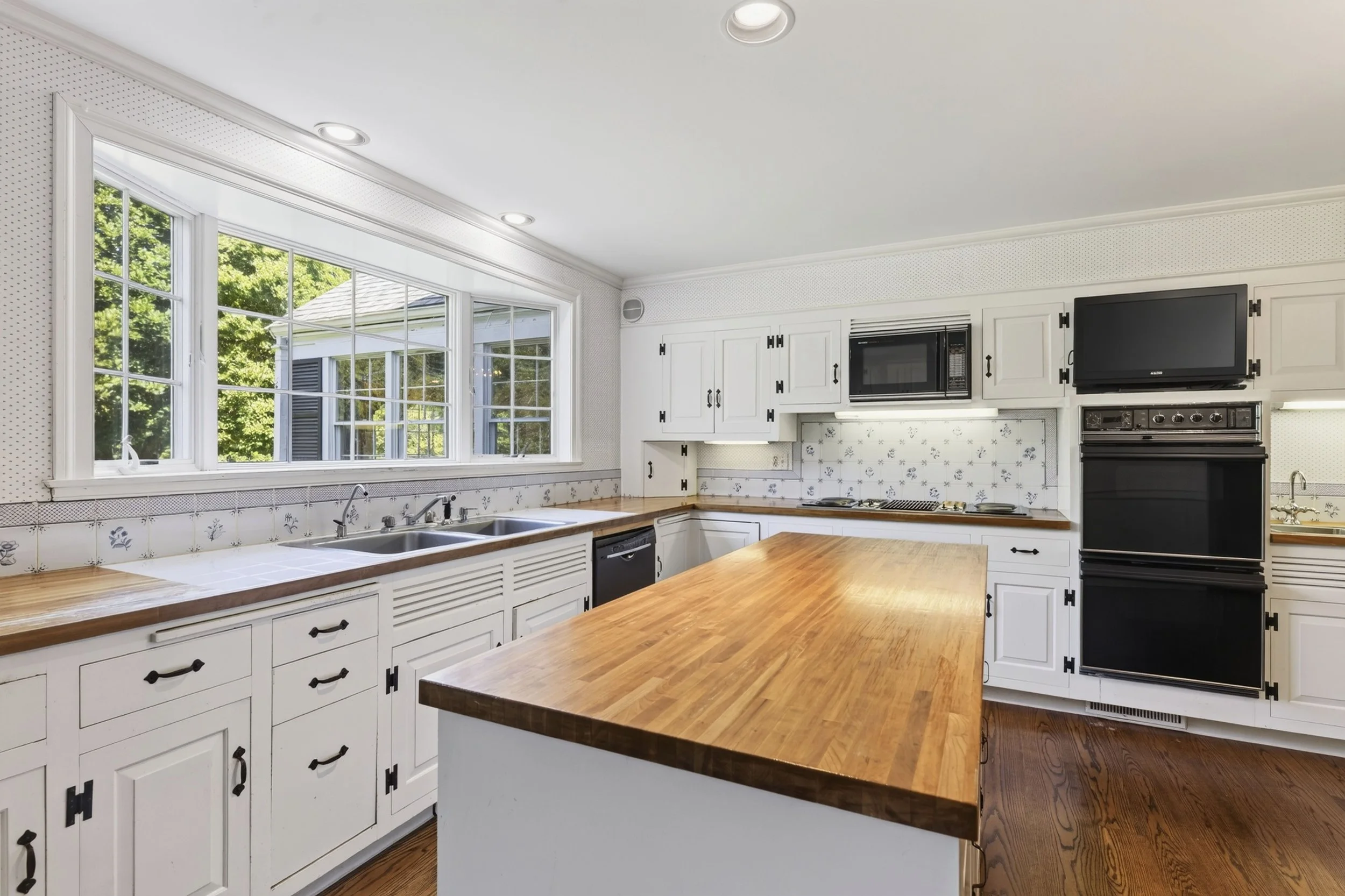 Bright kitchen with large window, white cabinets, black hardware, wooden countertops, black appliances including microwave and oven, and a wooden island.