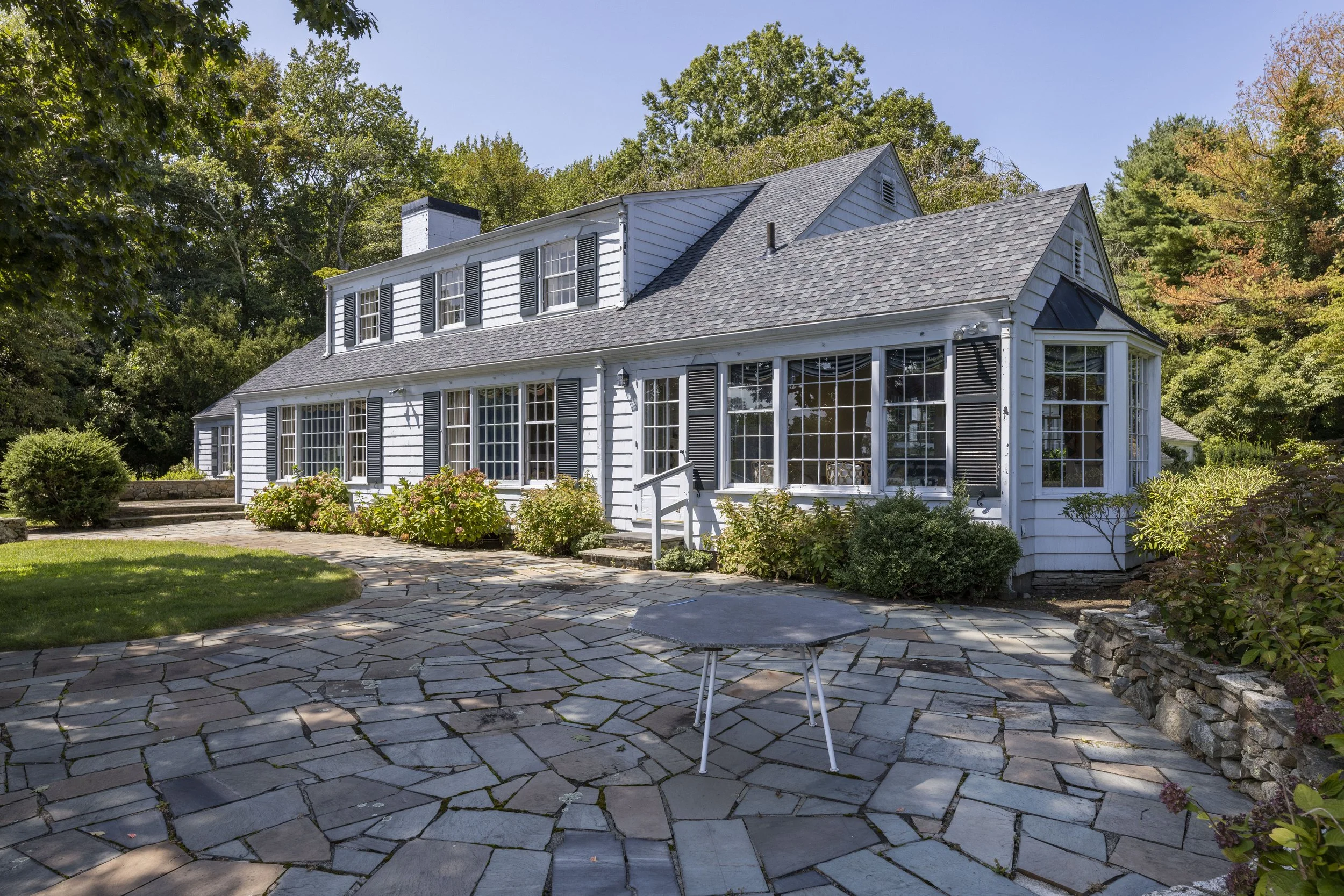A white house with black shutters and a screened porch, surrounded by greenery and a bistro table on a stone patio.