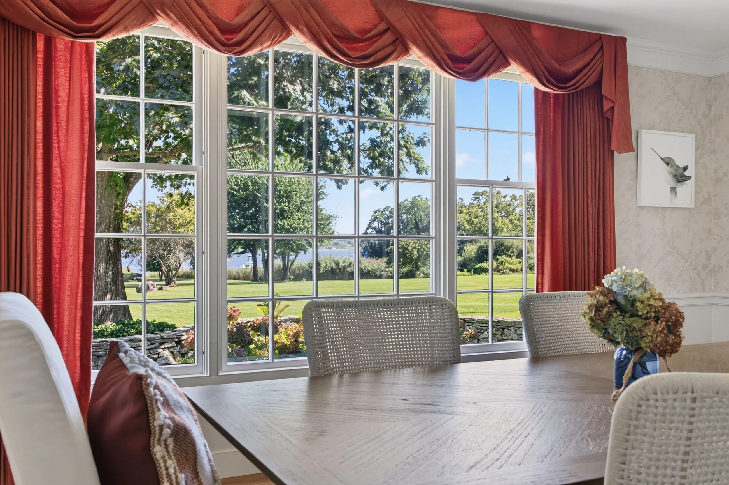 A dining room with large windows overlooking a green backyard with trees, a lake, and a blue sky. The windows are framed with red curtains and a valance. There is a table with a floral centerpiece.