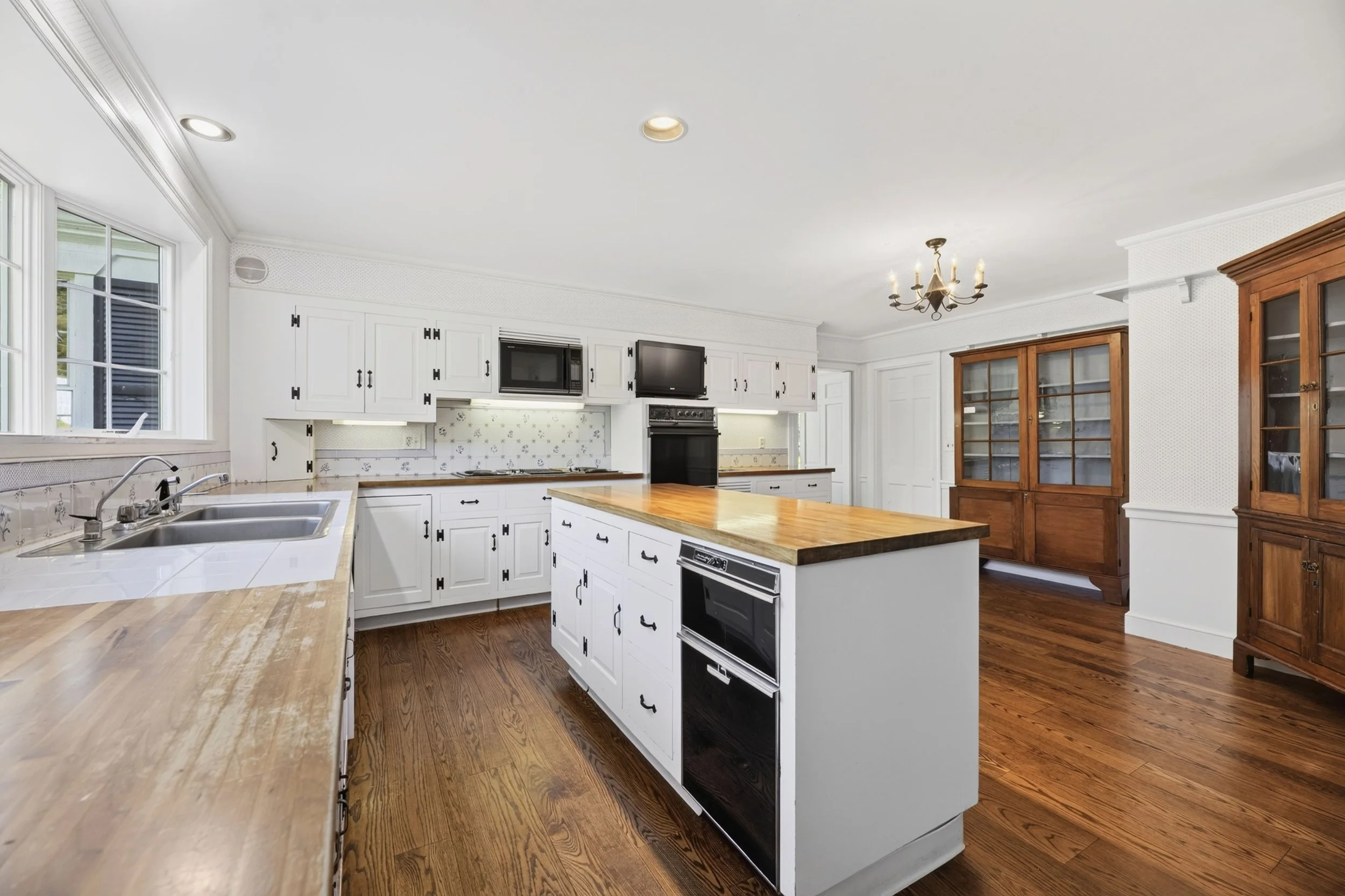 Bright kitchen with white cabinets, wooden countertops, a kitchen island, a wooden china cabinet, and a chandelier hanging from the ceiling.