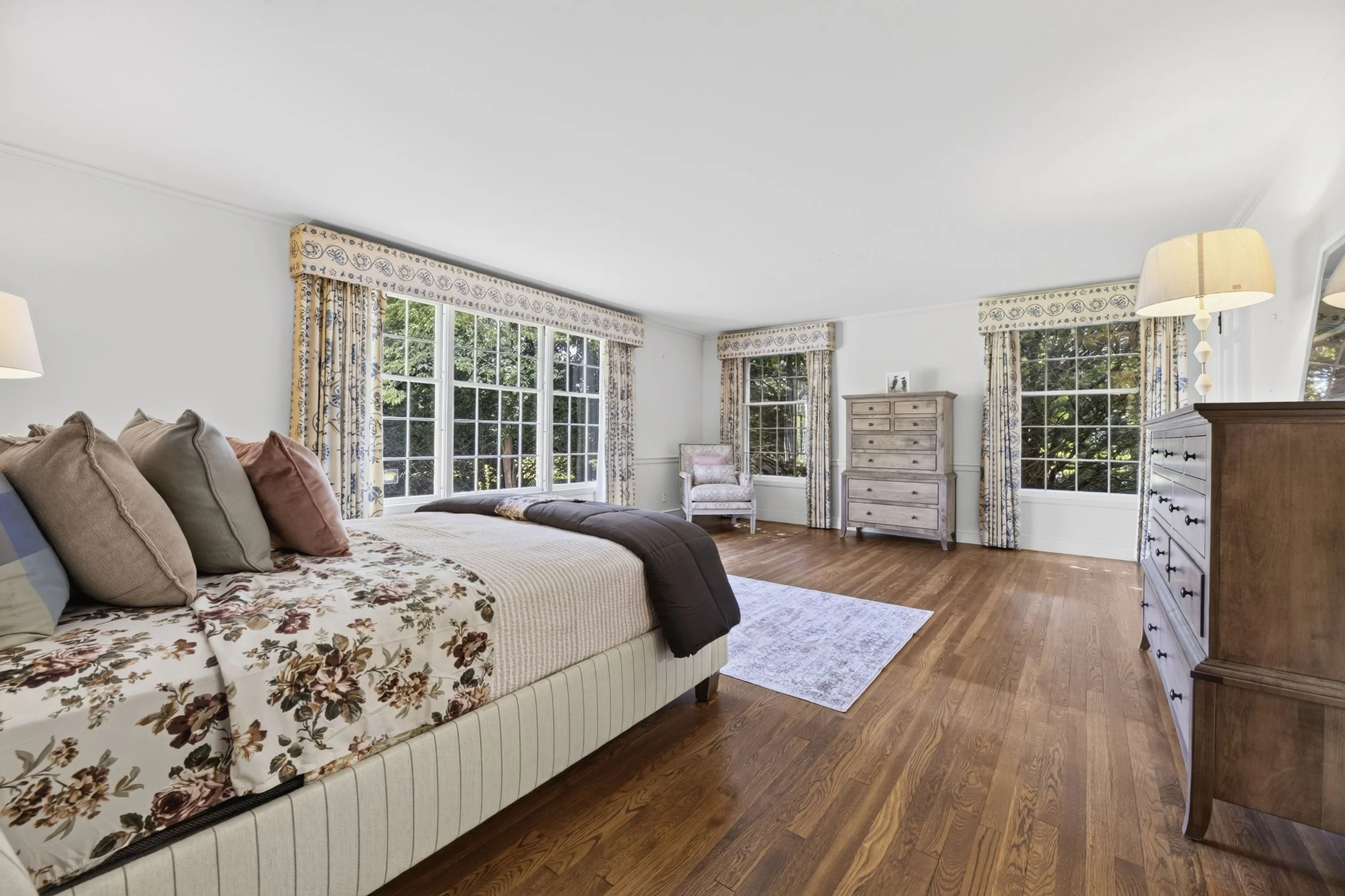 A bedroom with large windows, a bed with floral and beige bedding, wooden dresser and chest of drawers, an armchair, and beige curtains with a blue and yellow pattern.