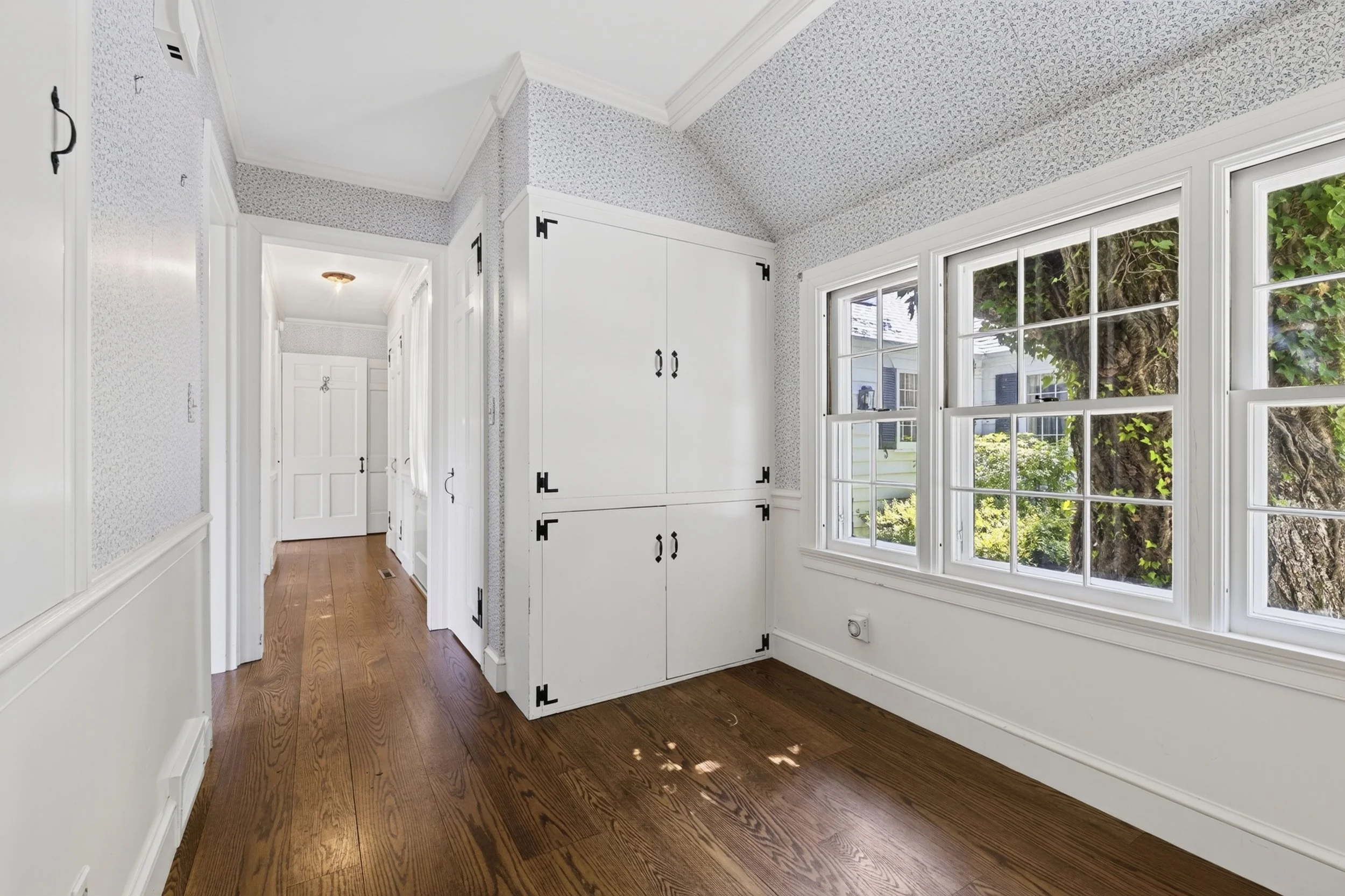 Interior of a house with large window, white walls with wallpaper, hardwood floors, and built-in cabinets, showing a hallway leading to a door.